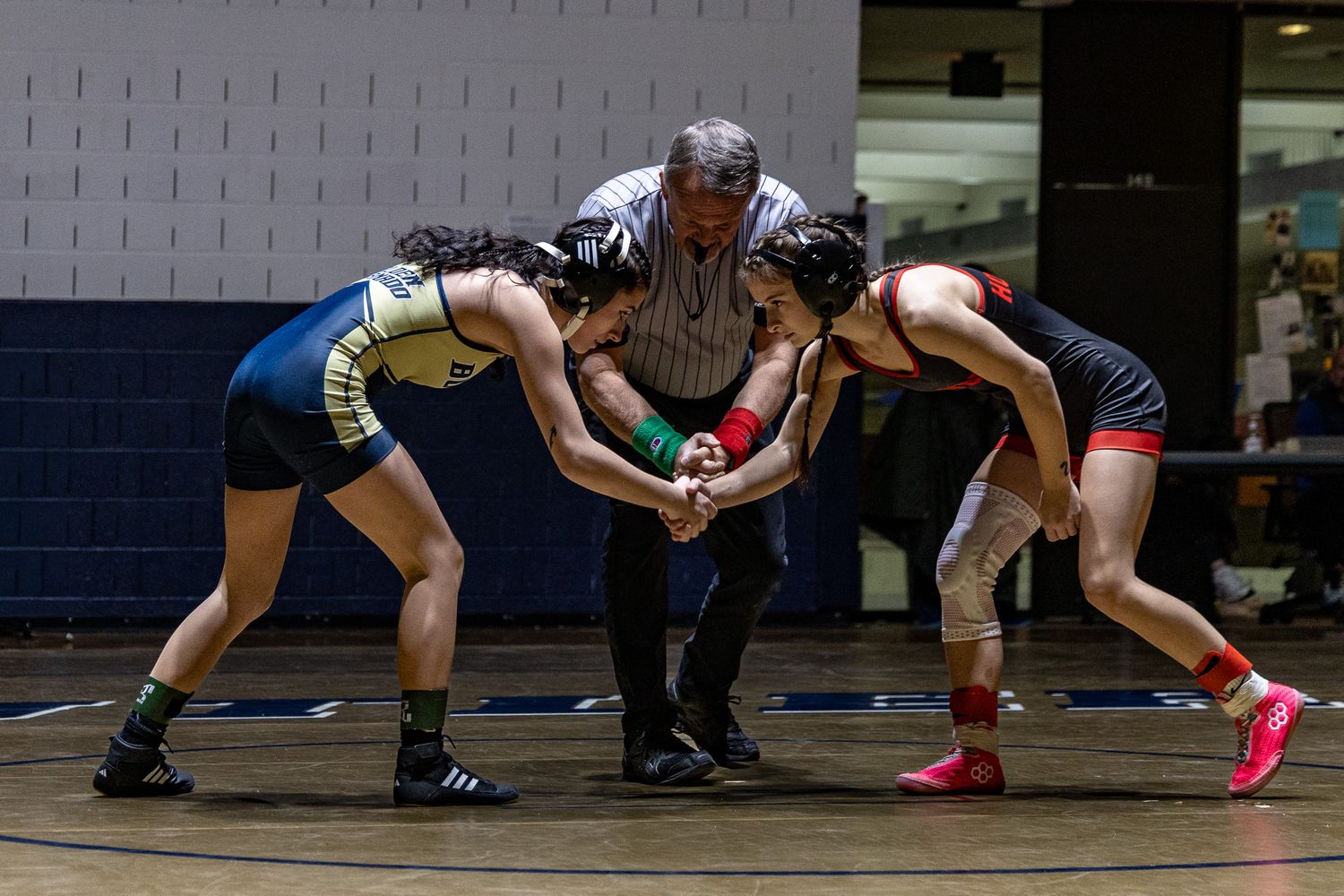 Referee oversees two female wrestlers shaking hands before match on blue gymnasium mat.