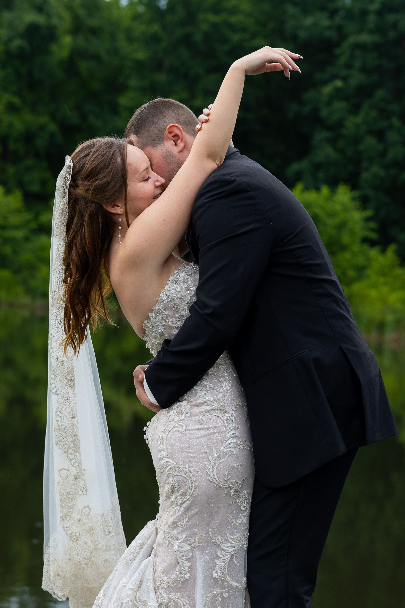 Bride and groom sharing passionate kiss by a lakeside, bride in lace mermaid gown with veil.