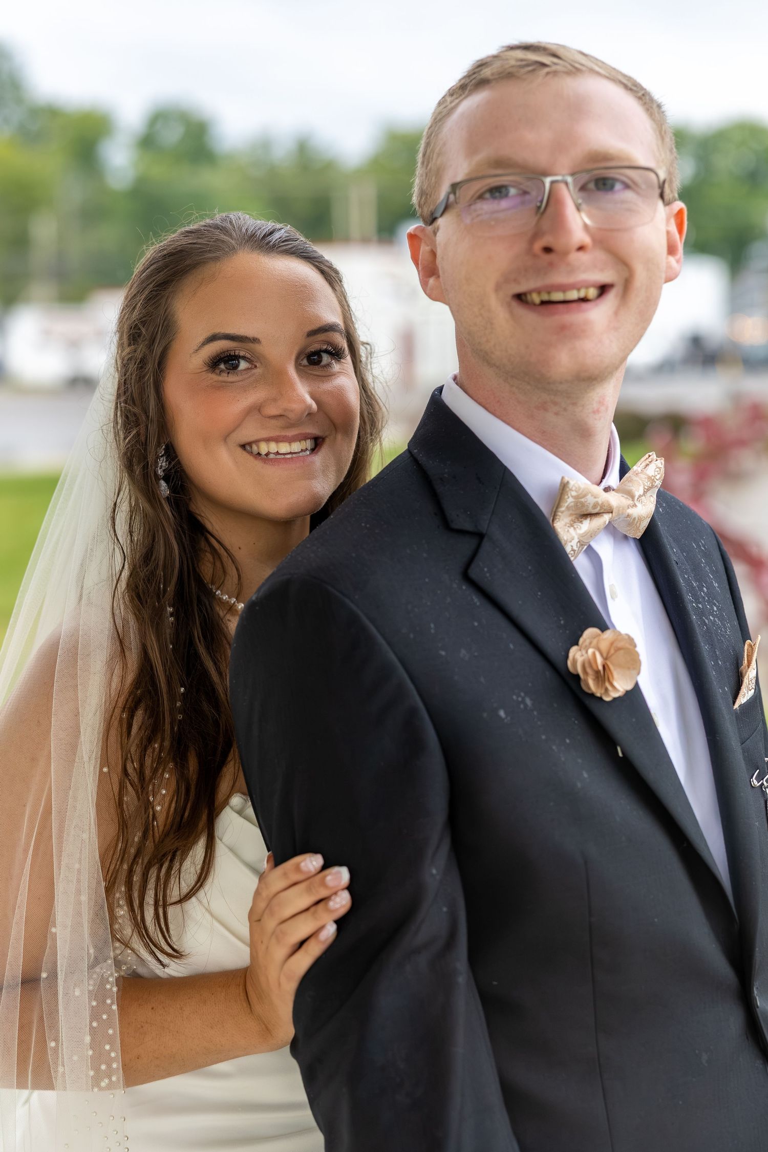 Smiling bride and groom posing outdoors on their wedding day, bride in white veil, groom in dark suit with bow tie.