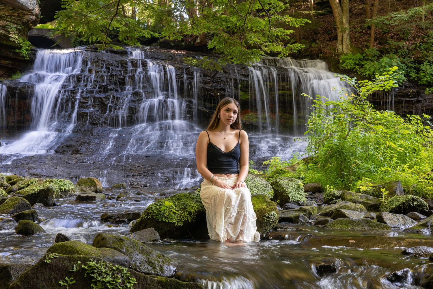Woman in black top and white skirt sits on rocks before a lush waterfall in a forest.