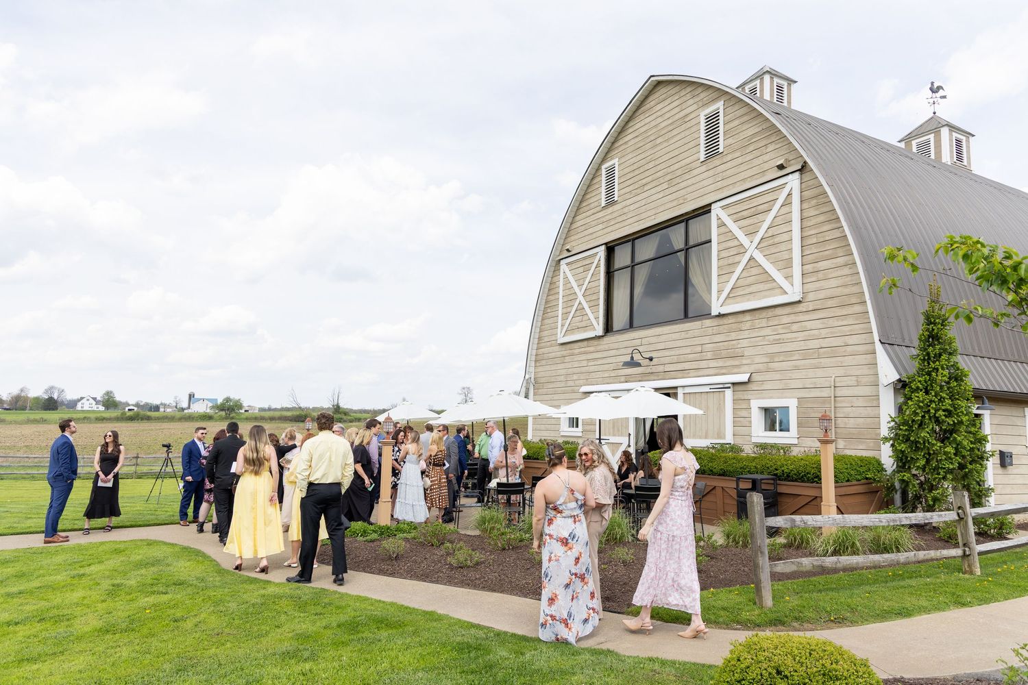 Wedding guests mingle outside a charming white barn venue surrounded by green lawns under a cloudy sky.