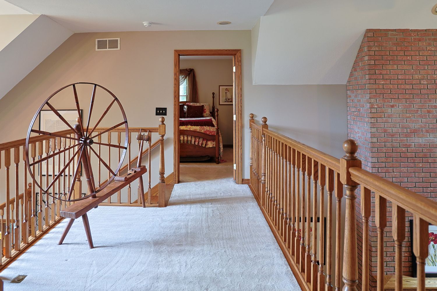 Upstairs hallway with antique spinning wheel, wooden railing, carpeted stairs, brick chimney, and bedroom doorway in a classic home.