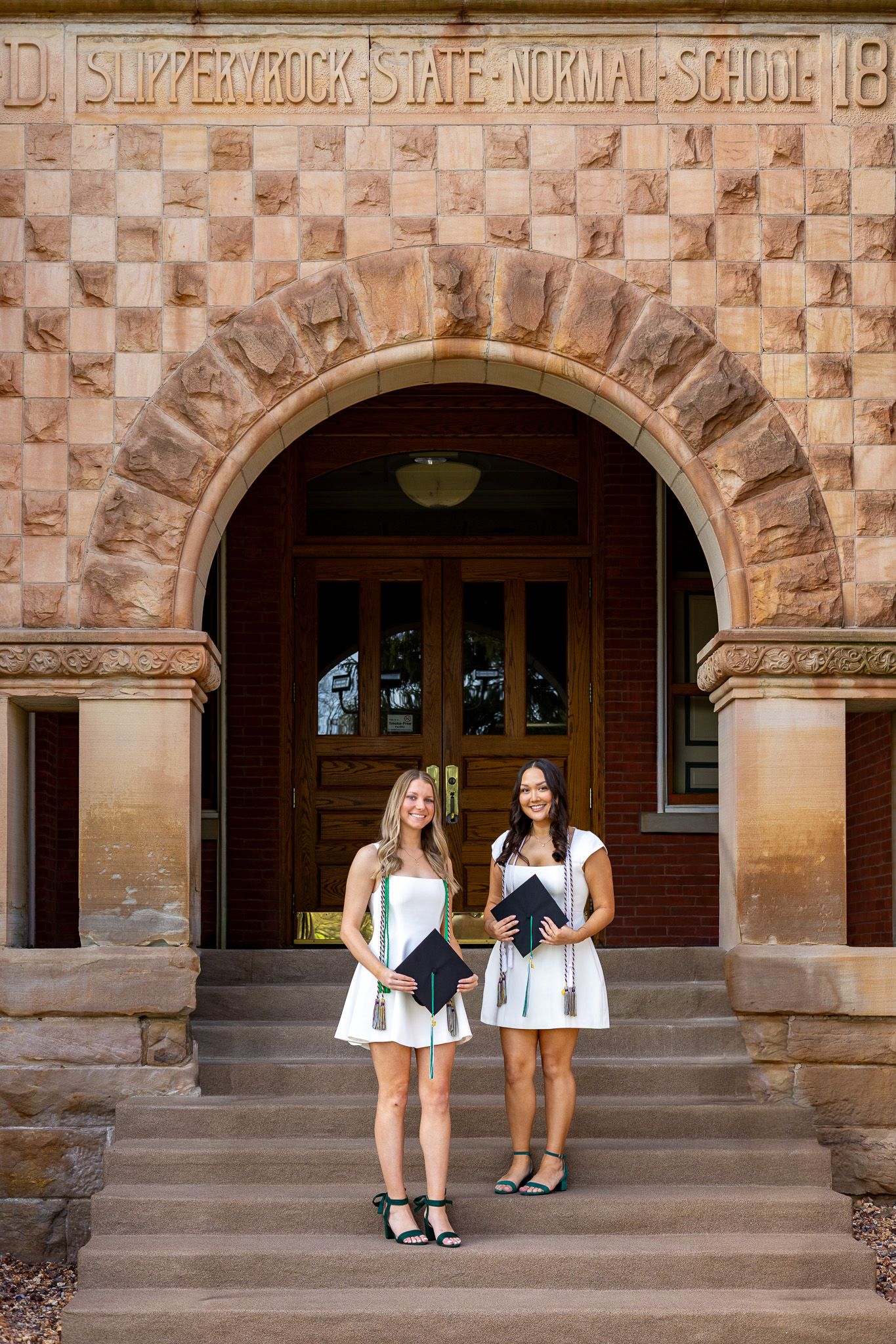 Two female graduates in white dresses hold caps on steps of Slippery Rock State Normal School.