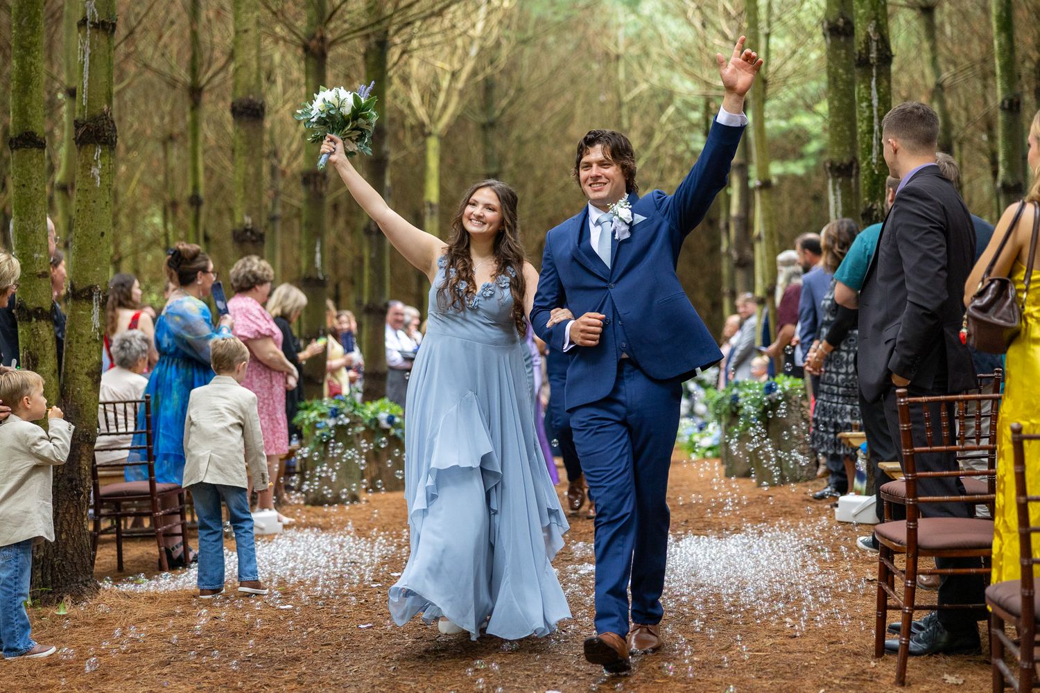 Newlywed couple celebrates joyfully while walking down a forest aisle lined with wedding guests.