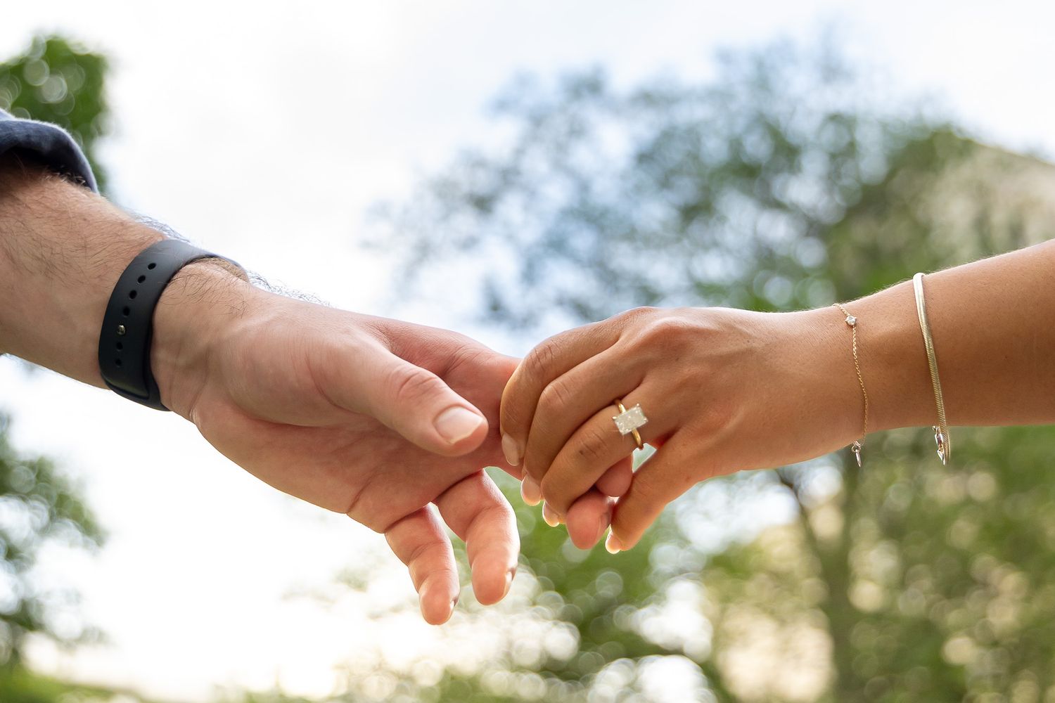 Close-up of couple holding hands outdoors, woman wearing diamond engagement ring and gold bracelet.