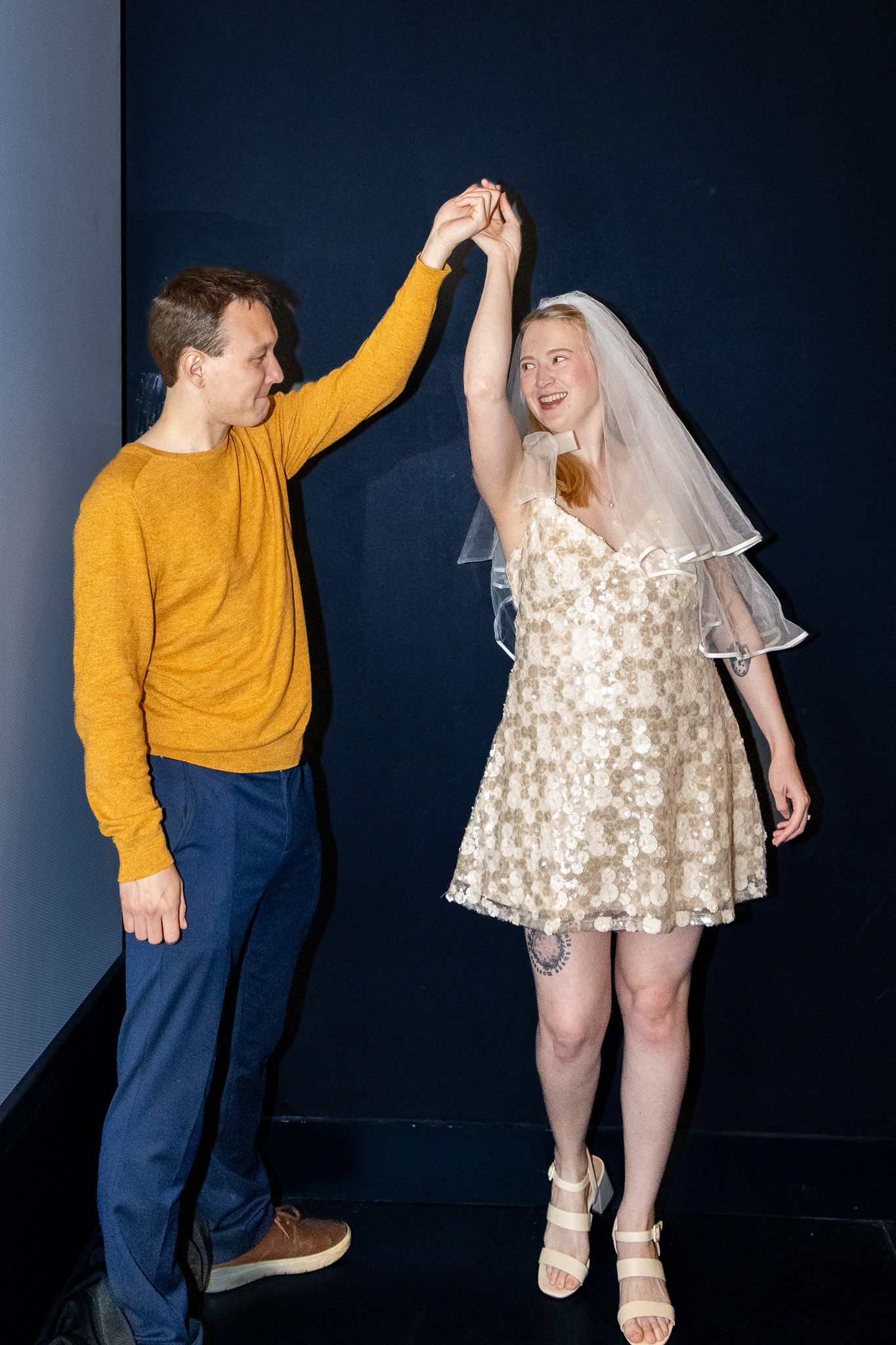 Couple dancing together, man in yellow sweater spinning bride in white mini dress and veil against dark background.