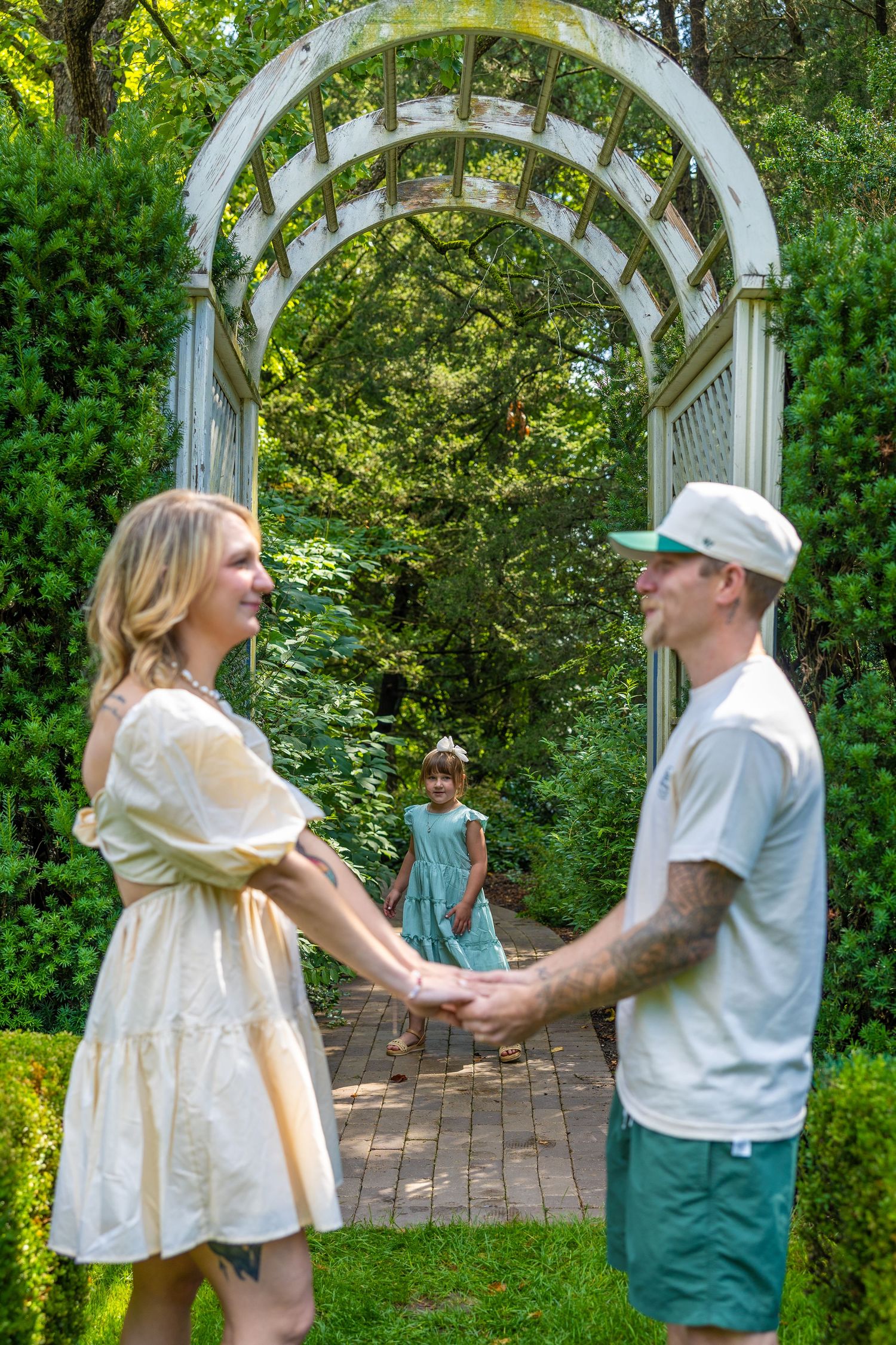 A couple holds hands in a lush garden archway while a young girl stands behind them on a brick path.