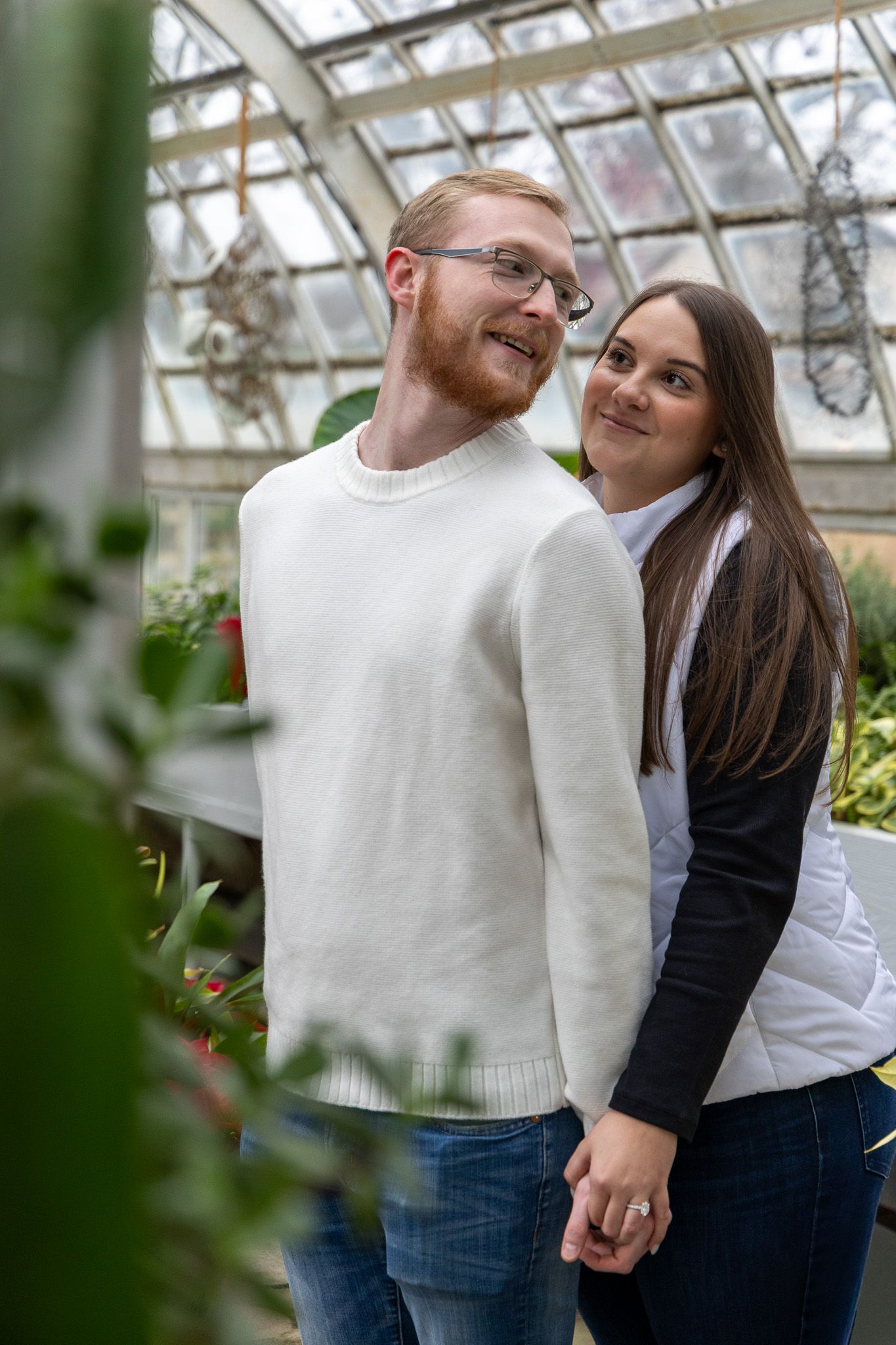 A couple holding hands and smiling in a greenhouse, man in white sweater with red beard, woman in black and white vest.