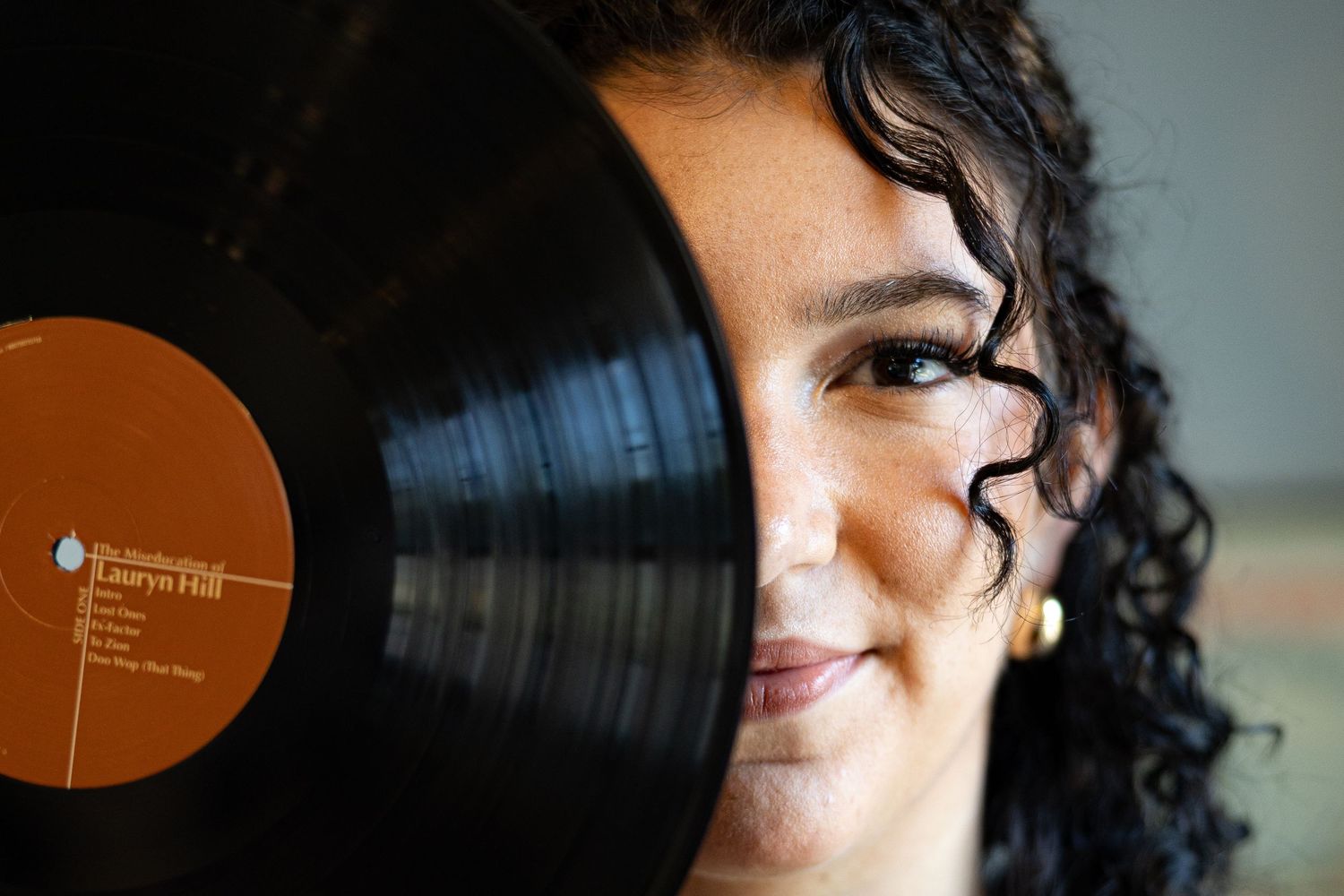 Woman smiling behind a Lauryn Hill vinyl record, showing one eye and a gold earring.