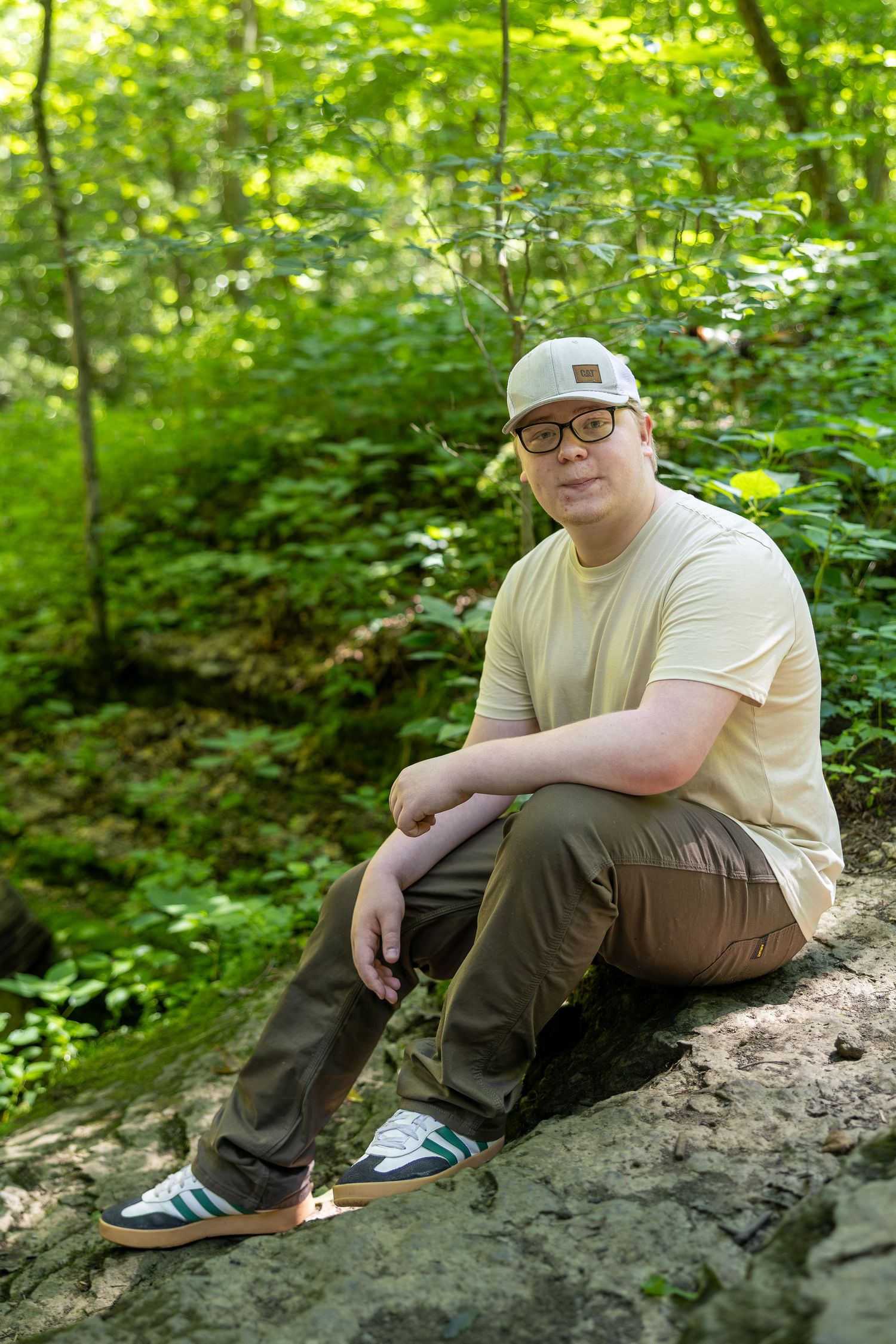 Man in cap and glasses sitting on a rock in a lush green forest wearing olive pants and a yellow t-shirt.