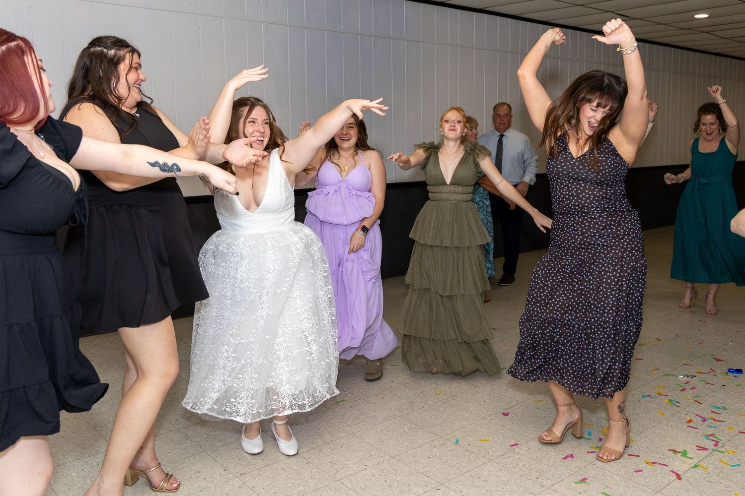 Bride and guests dancing joyfully at wedding reception, wearing formal dresses in white, purple, and black polka dot.