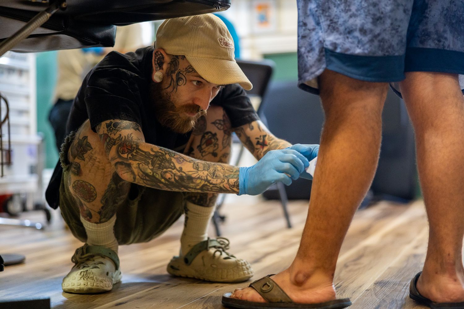 A heavily tattooed artist carefully applies a tattoo to a client's foot in a colorful studio.