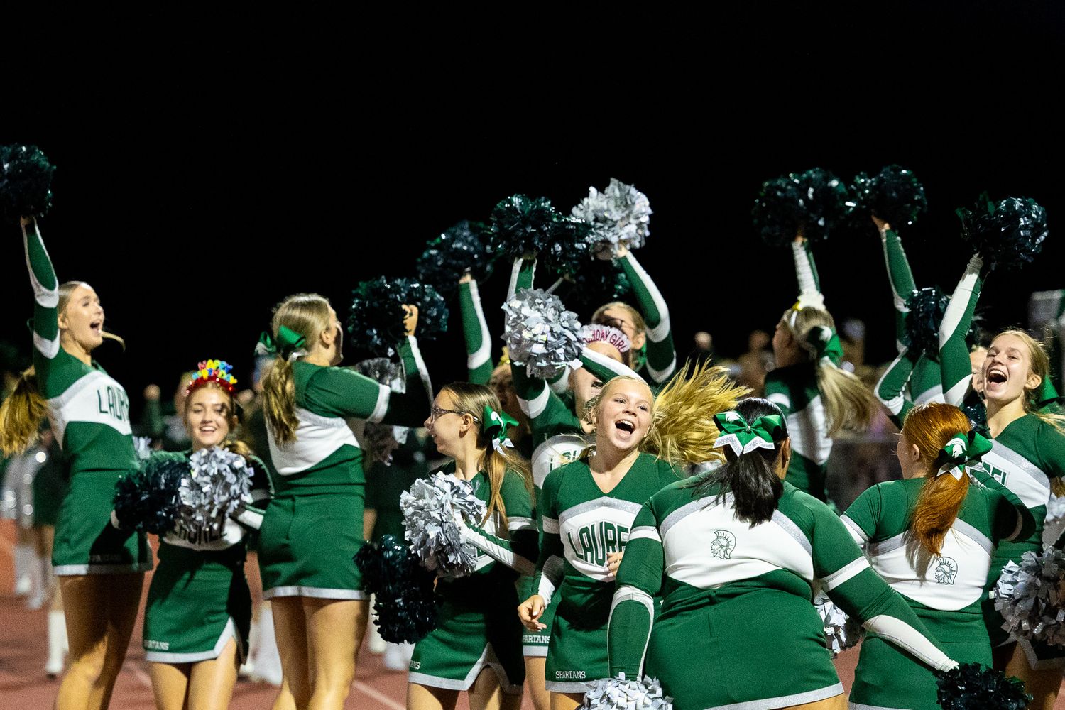 Energetic cheerleaders in green and white uniforms wave silver pom-poms during a nighttime football game.