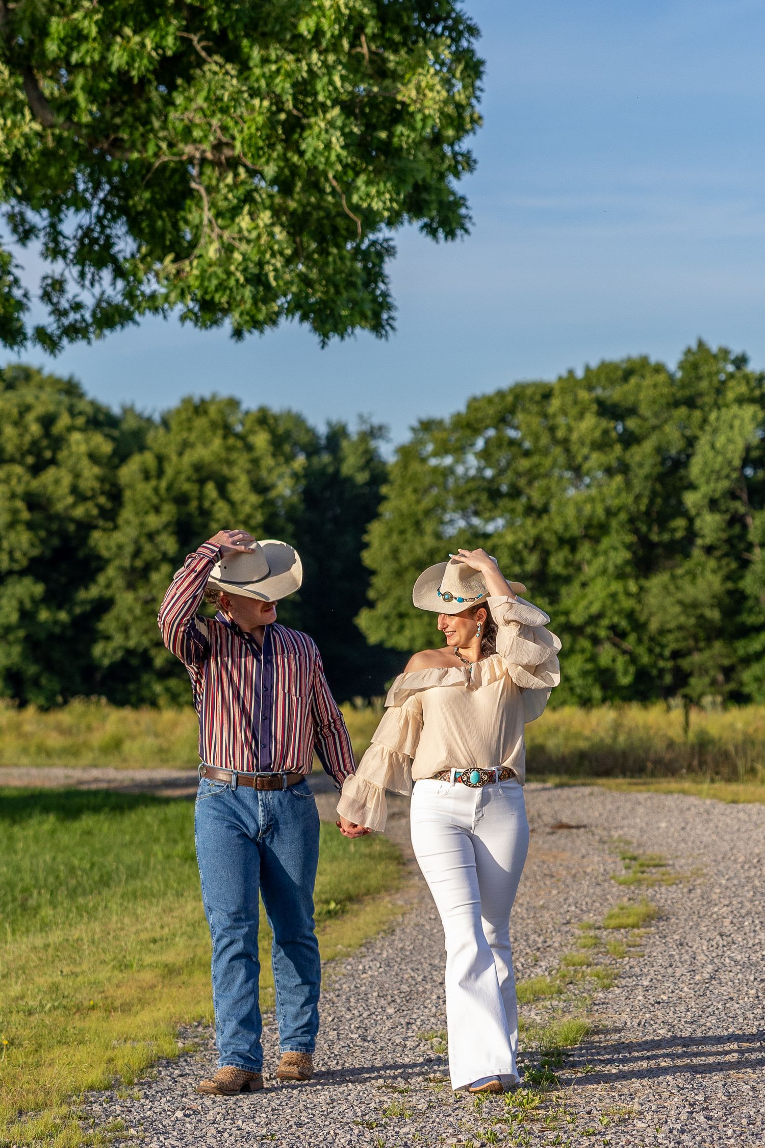 A couple in cowboy hats holds hands while walking down a sunny gravel road lined with green trees.