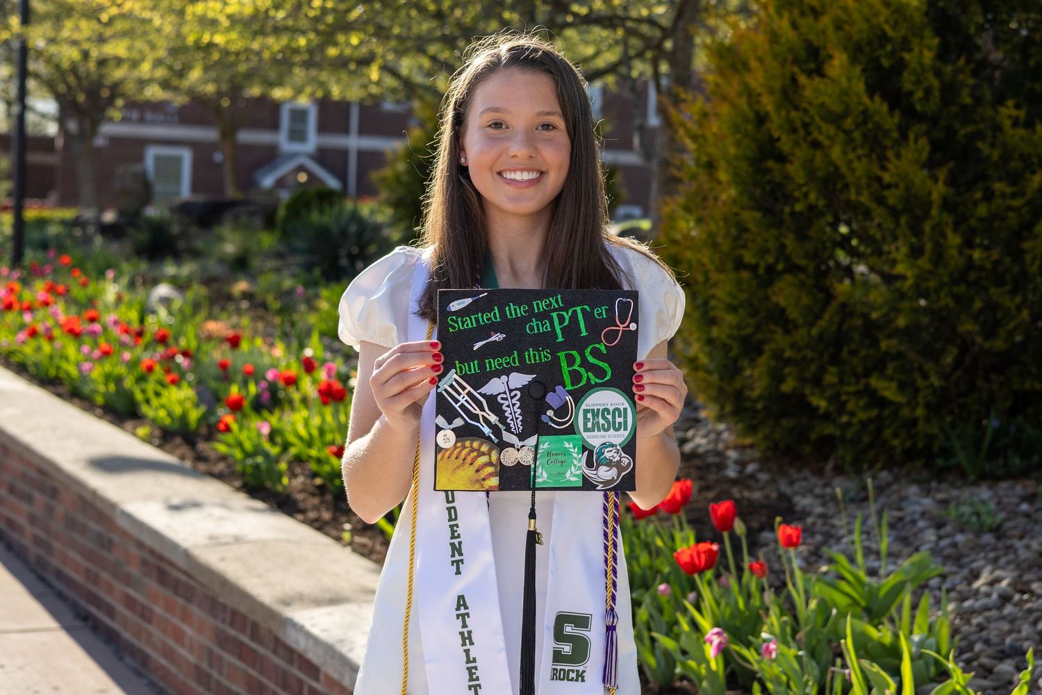 Smiling graduate holds decorated cap reading PT BS beside colorful tulips at Slippery Rock University.