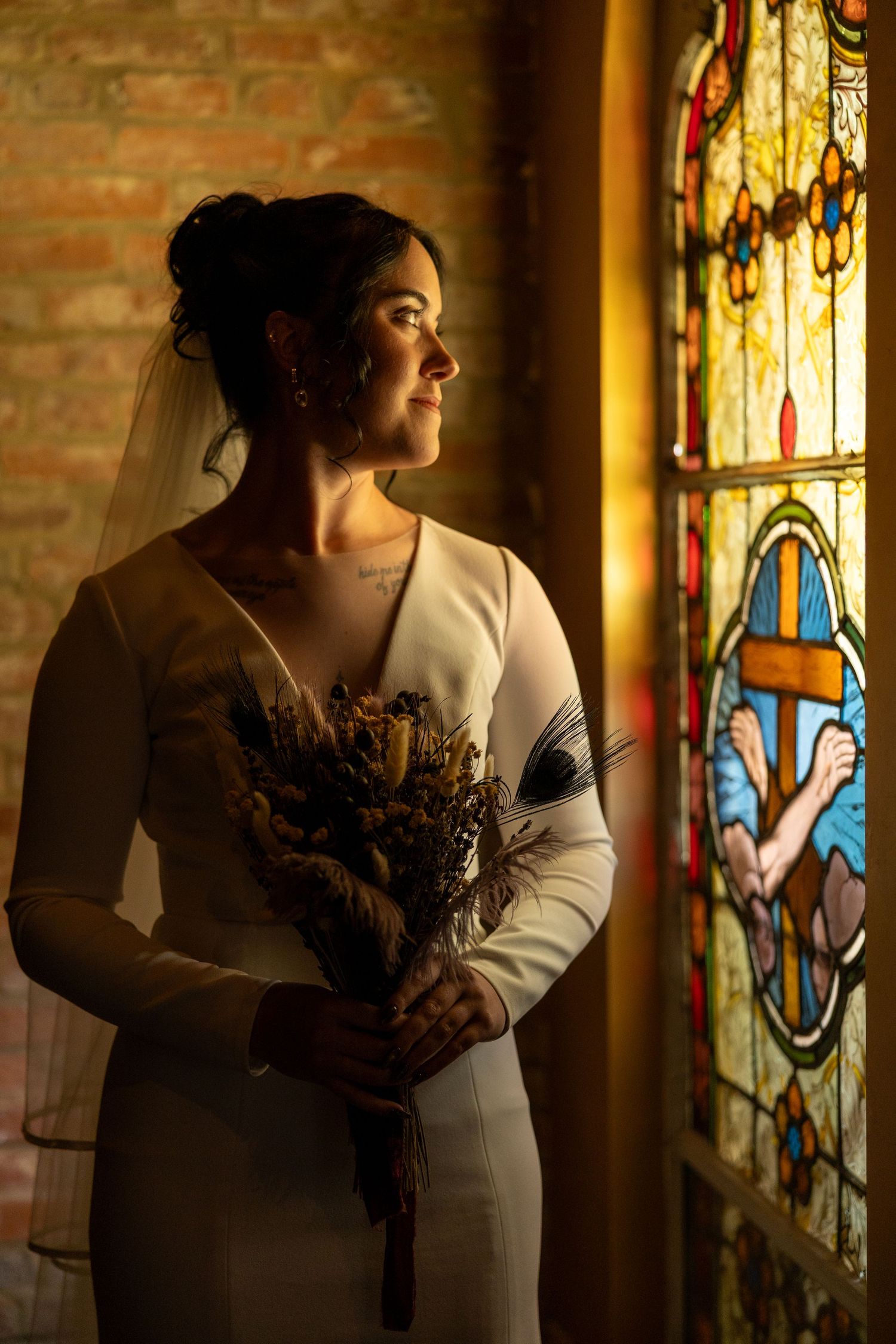 Bride in white dress holding dried bouquet stands by stained glass window in warm church light.
