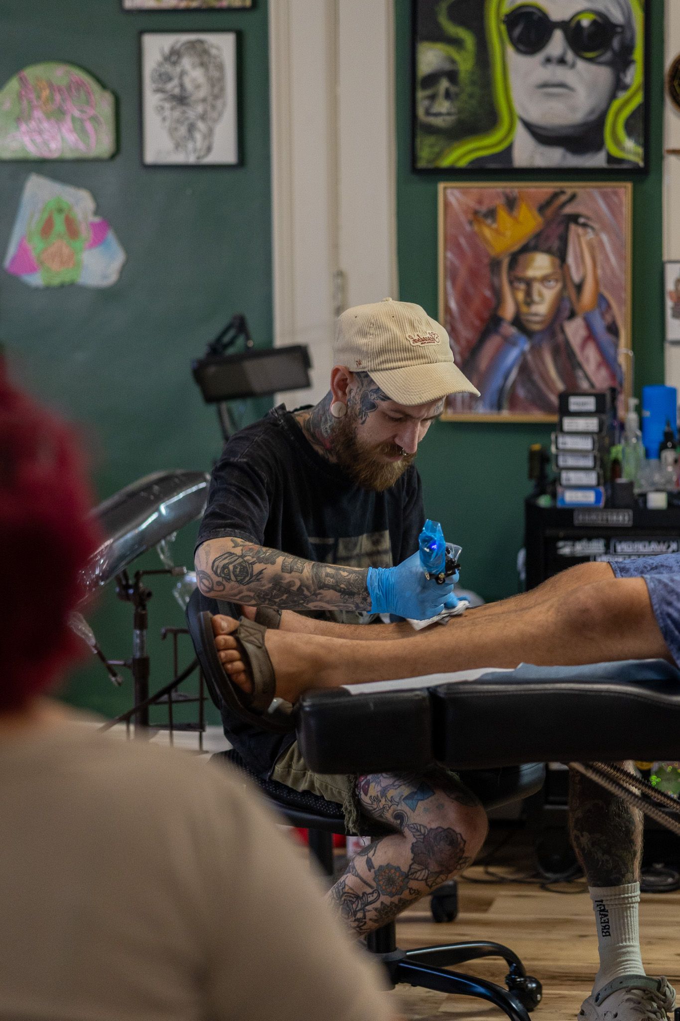 Bearded tattoo artist with cap and sleeve tattoos inking a client's leg in an art-filled studio.
