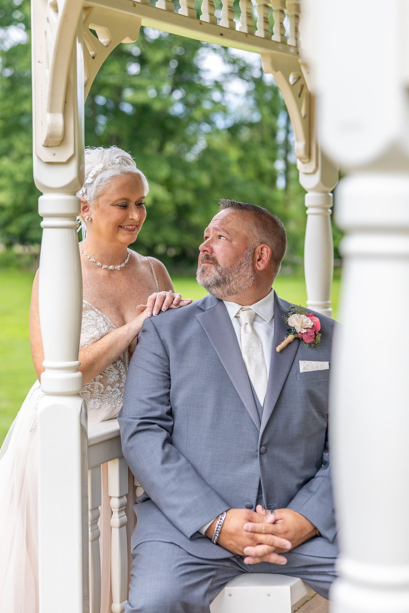 Bride and groom share a tender moment in a white gazebo, she in lace gown, he in gray suit with boutonniere.