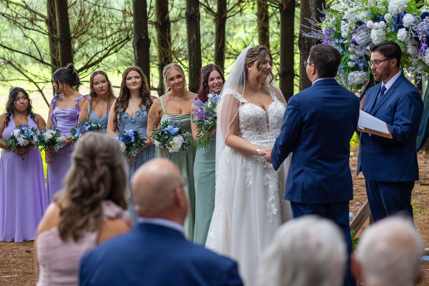 Bride exchanges vows with groom as bridesmaids in purple and green dresses look on in forest ceremony.