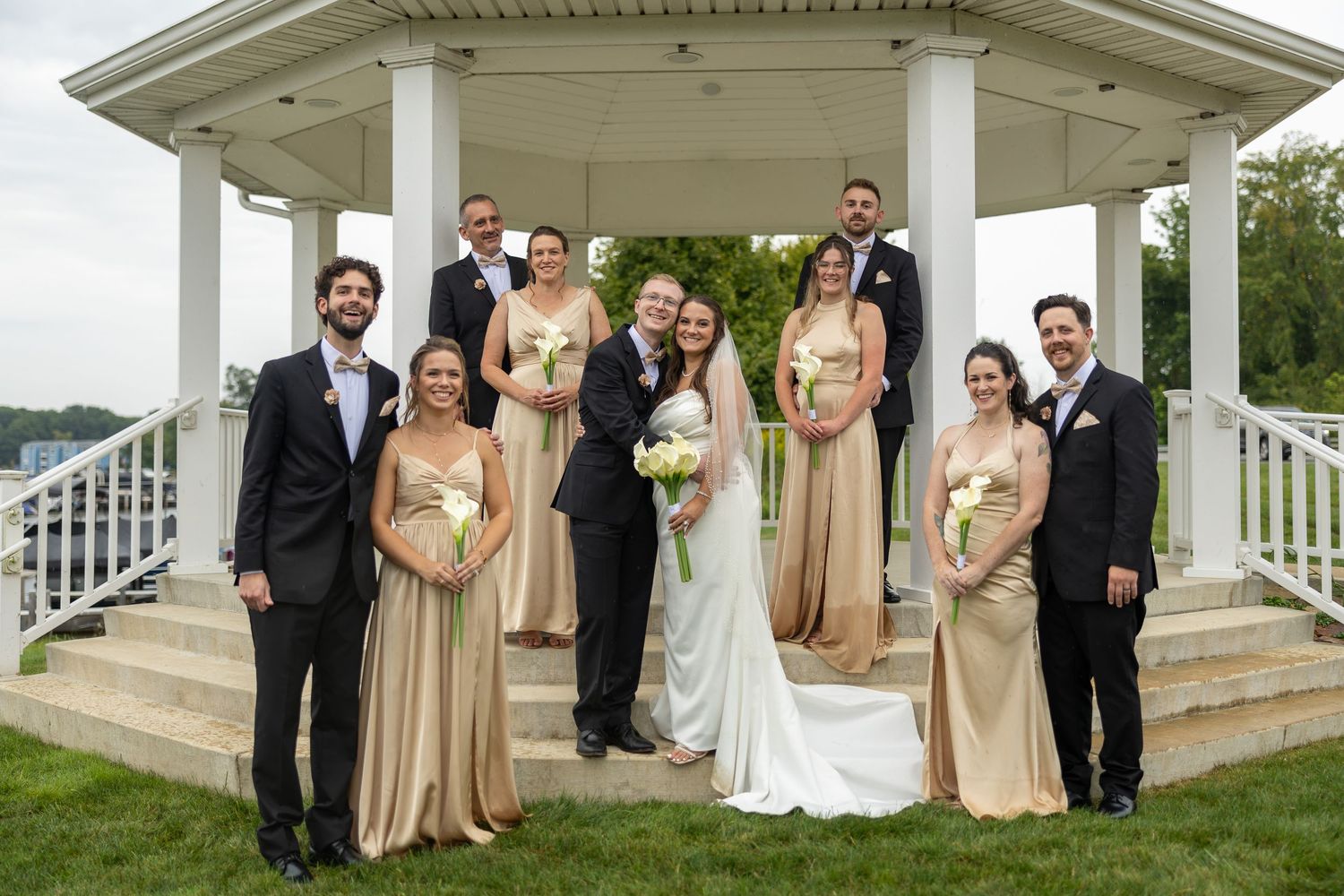 Wedding party poses in front of a white gazebo, bridesmaids in gold gowns and groomsmen in black tuxedos.
