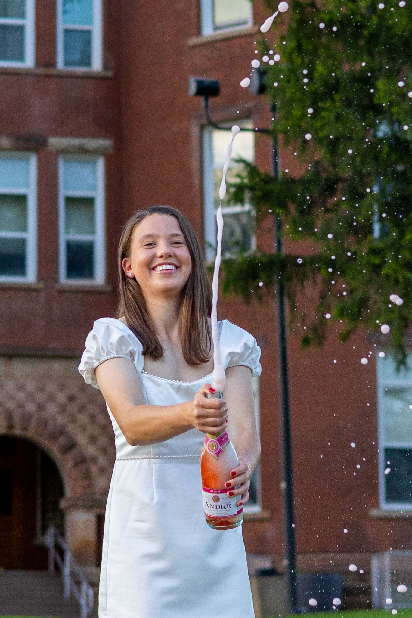 Smiling young woman in white dress popping a bottle of sparkling wine outside a red brick college building.