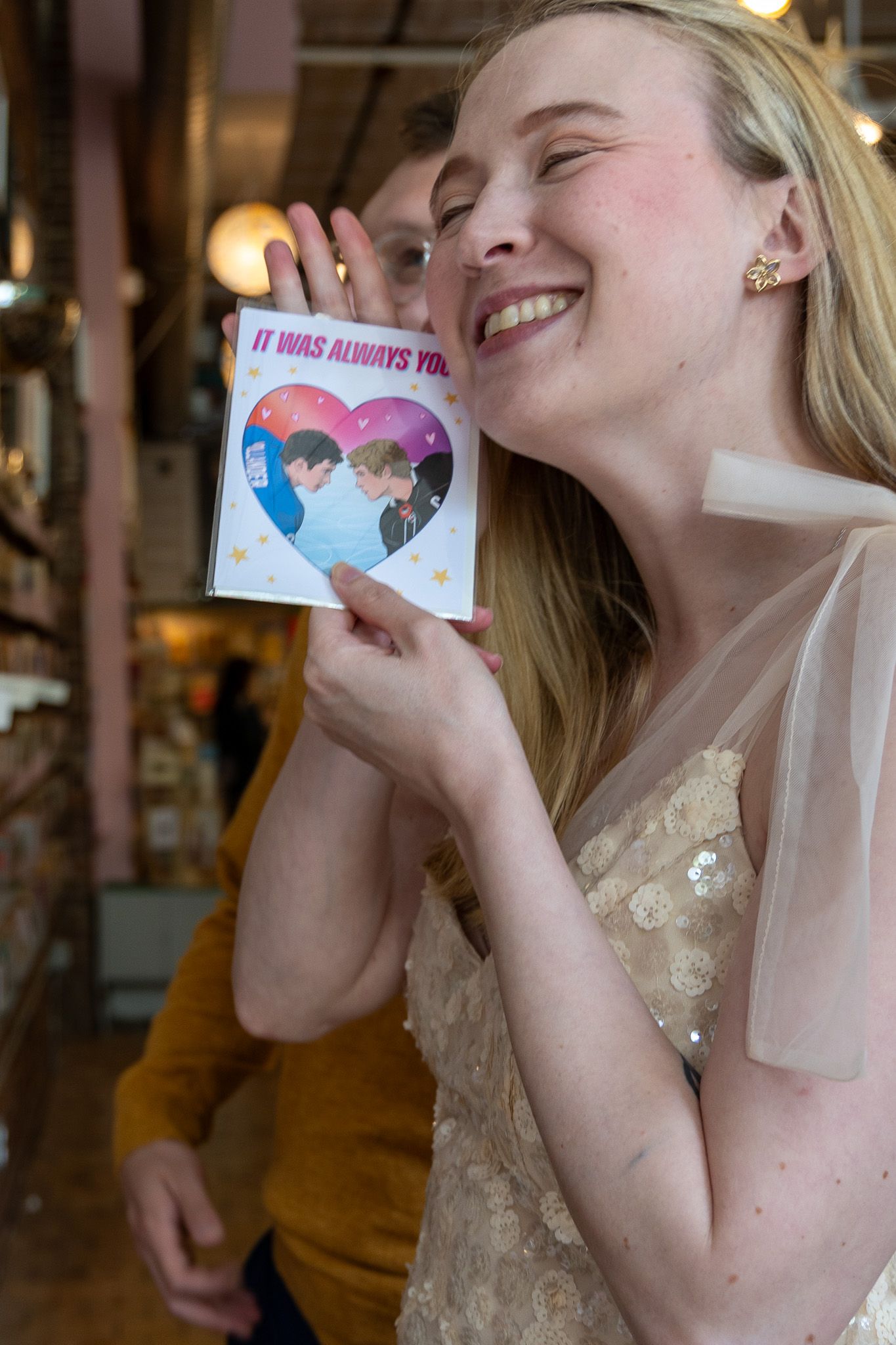 Smiling blonde woman holding heart-shaped card reading It Was Always You in a bookstore.