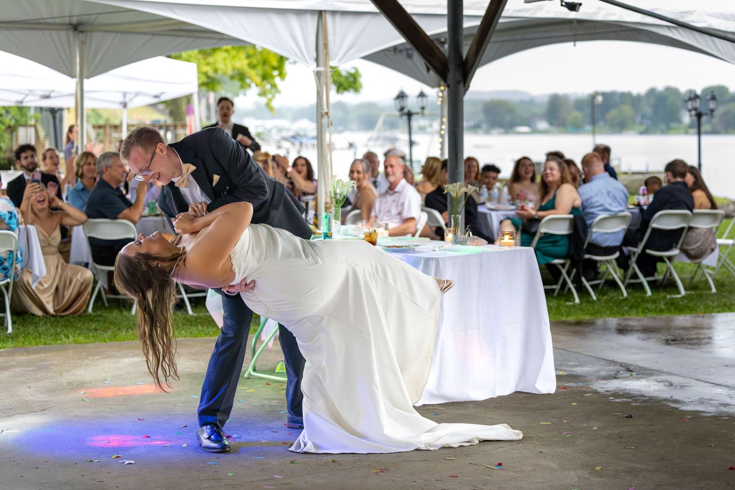 Bride and groom share romantic dip dance at outdoor waterfront wedding reception with guests watching.