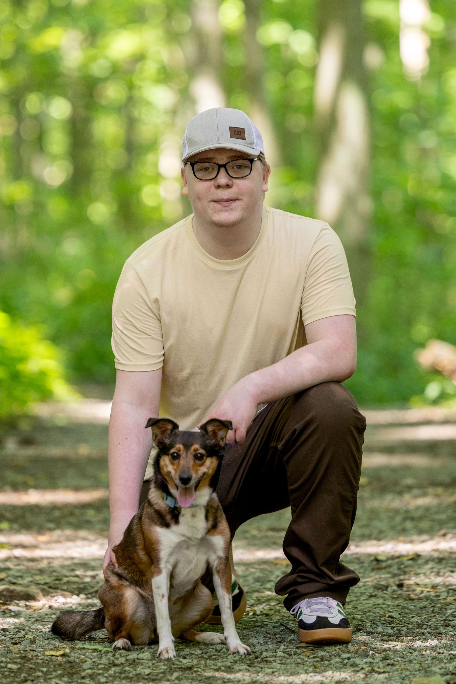 Young man in cap and glasses crouching with his dog on a forest path.