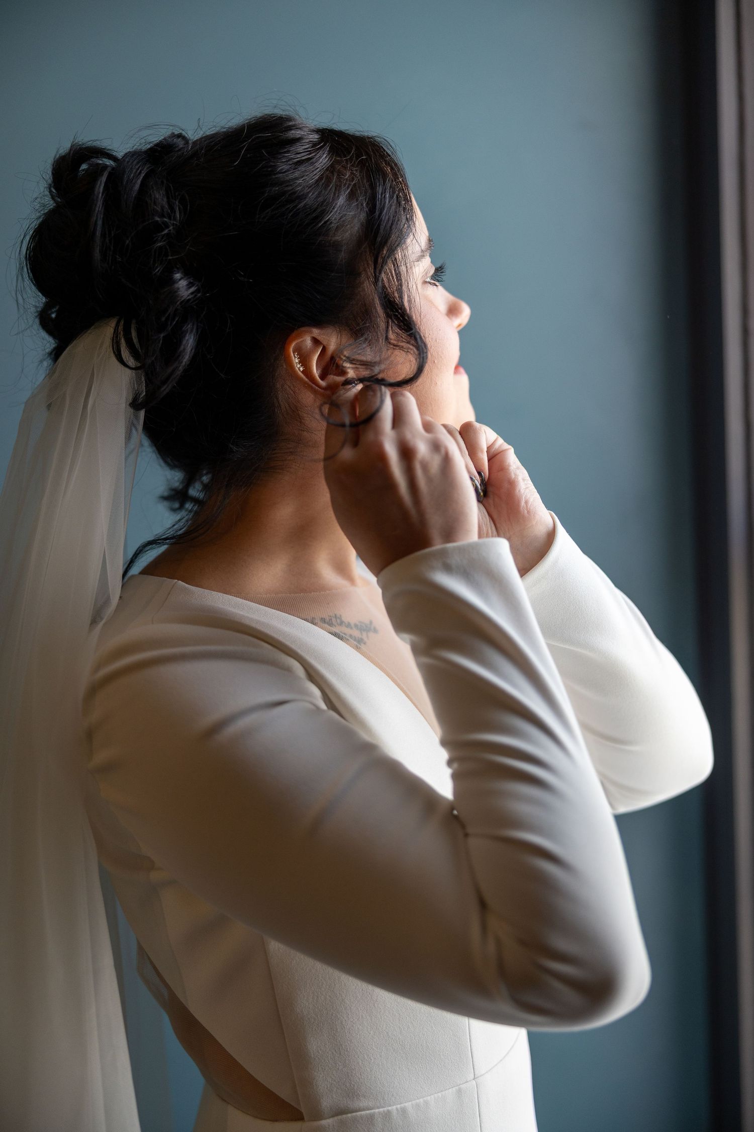 Bride in white long-sleeve gown adjusting earring near window, dark hair in updo with veil.