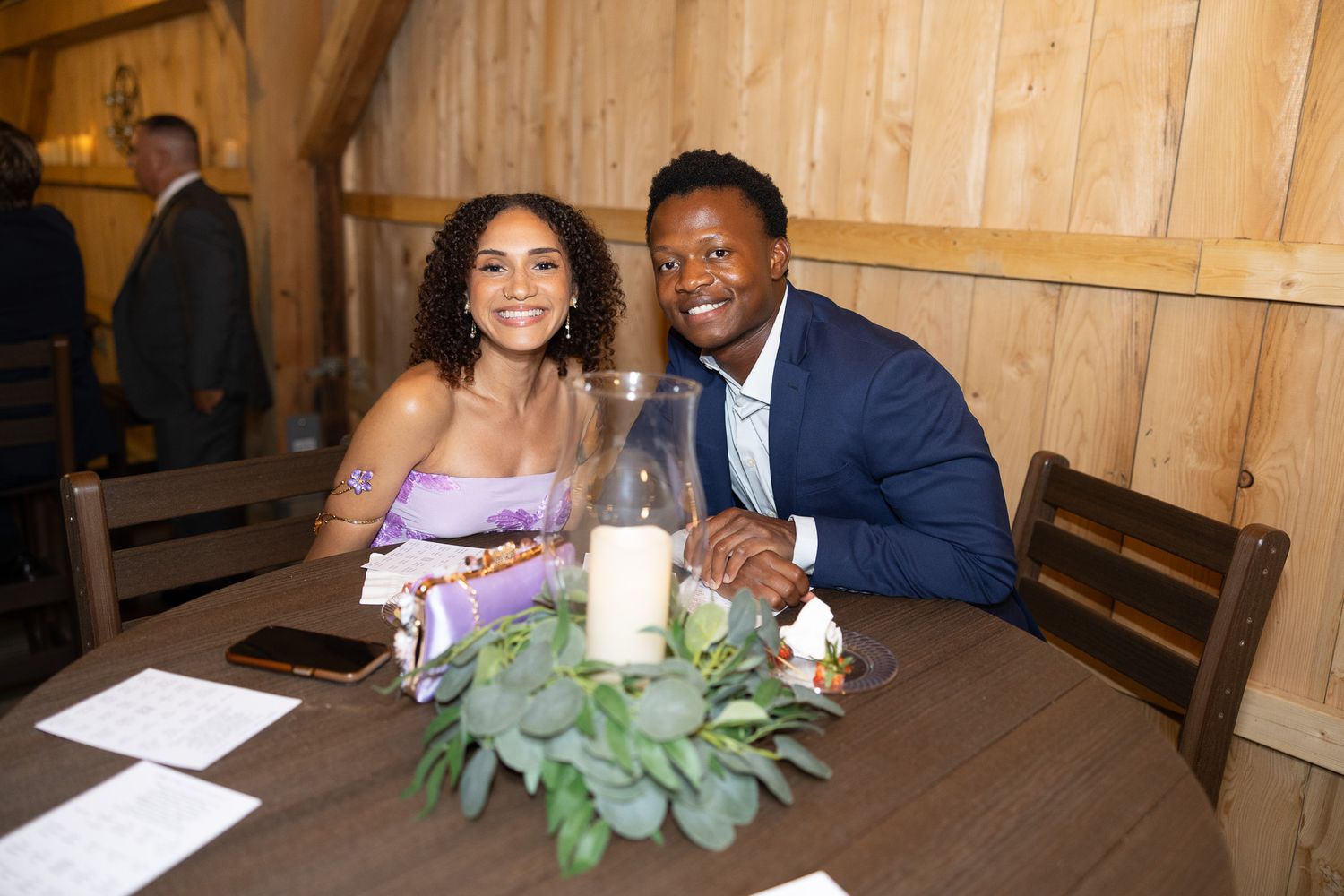 Couple smiles warmly at a wedding reception table adorned with greenery and candle centerpiece.