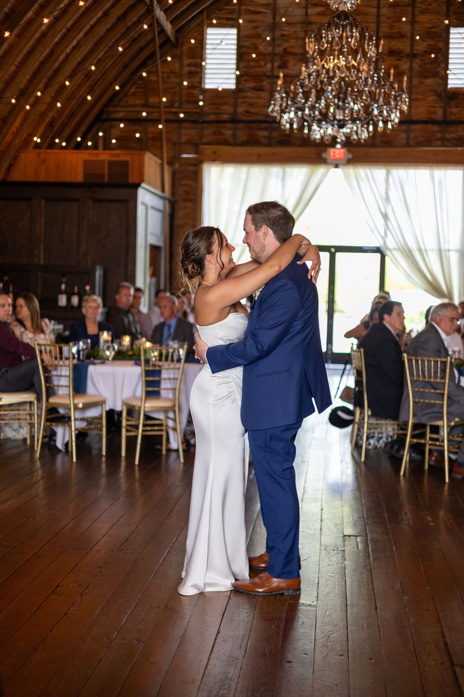 Bride and groom share their first dance in a rustic barn reception hall with chandeliers and string lights.