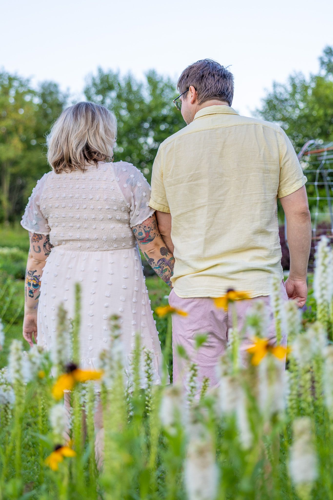 Couple with tattoos holding hands walking through a colorful wildflower garden from behind.