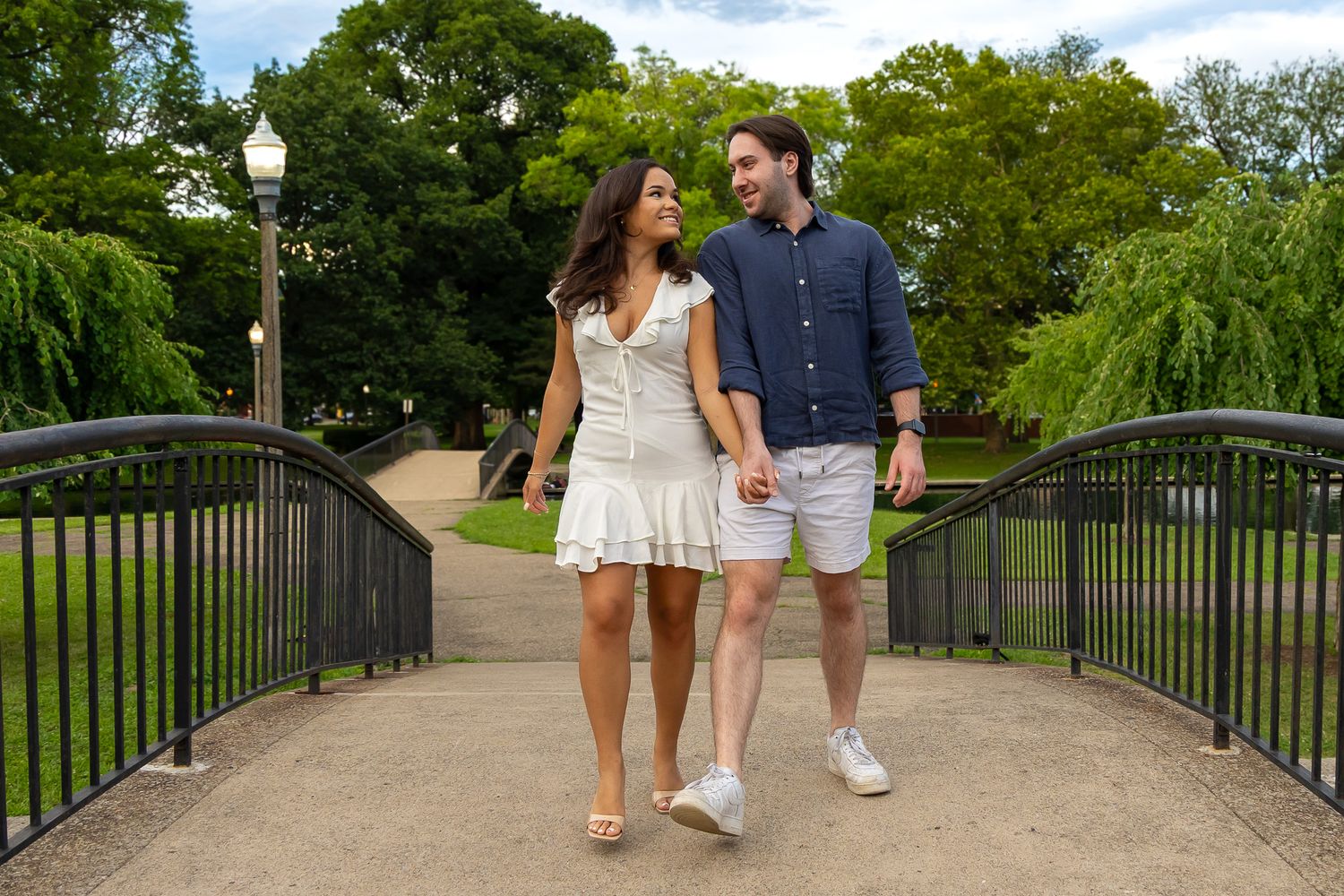 Happy couple walking hand in hand on a bridge in a lush green park during engagement session.