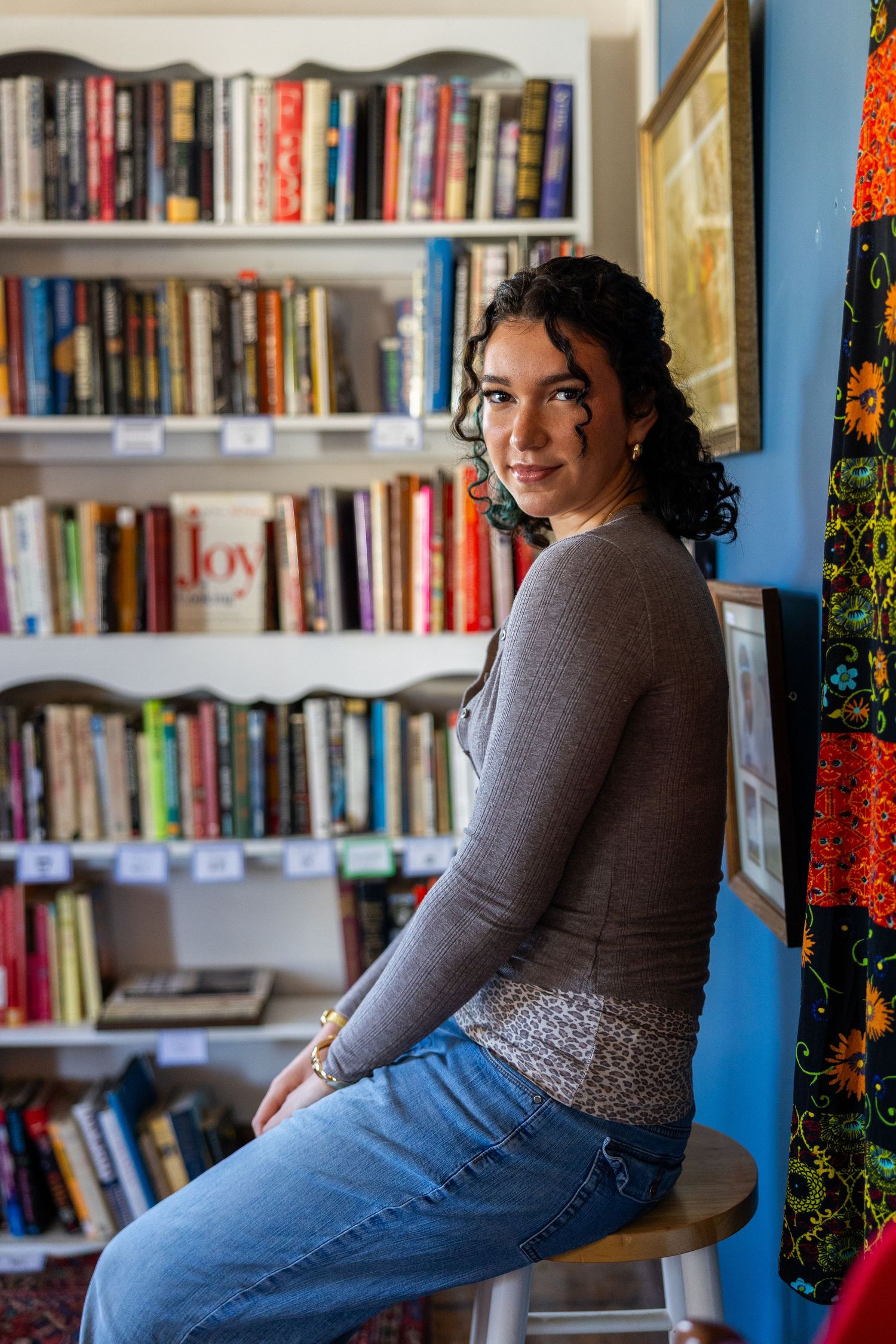 Young woman with curly hair sits on stool in front of colorful bookshelf in cozy home library.