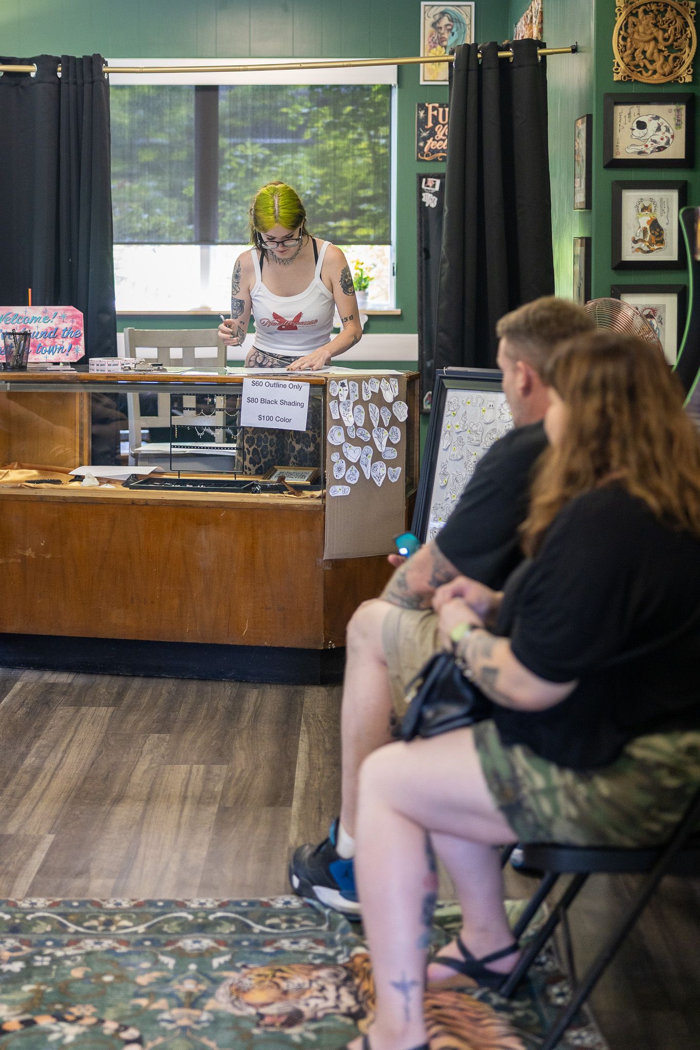 Two customers wait while a green-haired tattoo artist works at a reception desk in a decorated tattoo parlor.