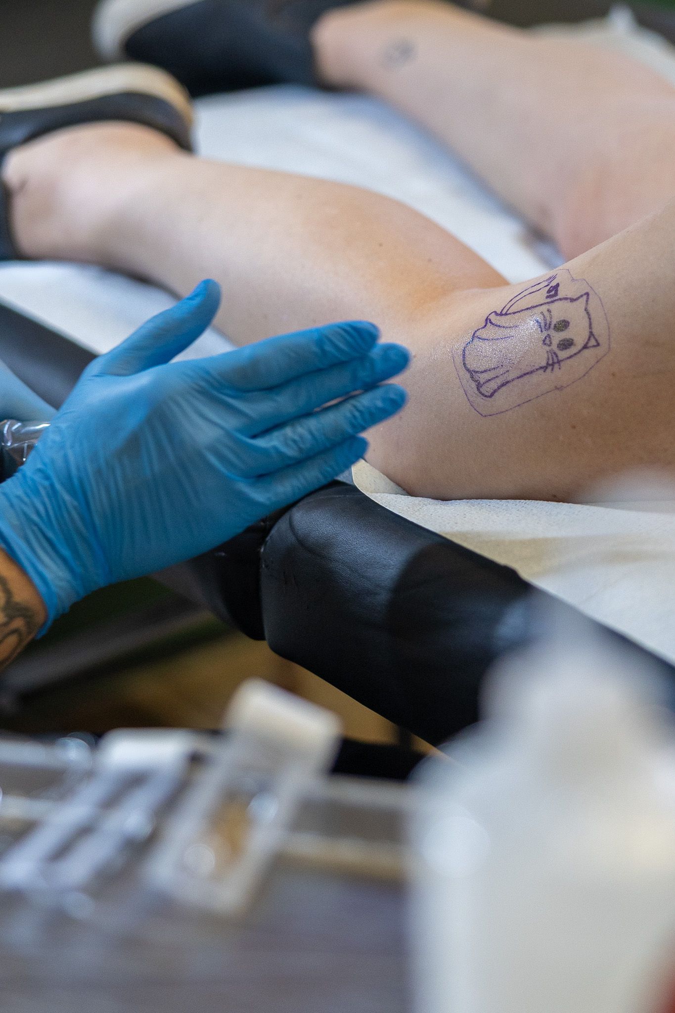 Tattoo artist in blue gloves steadies a clients arm showing a skull stencil outline ready to be tattooed.