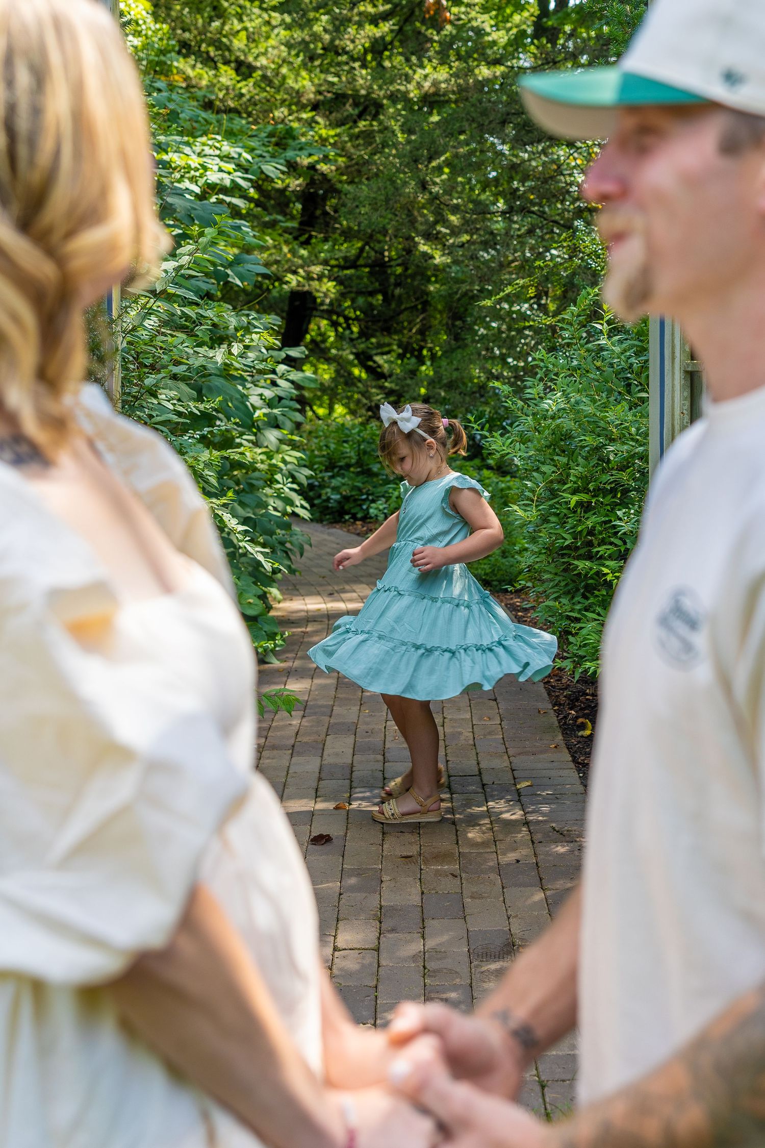 Young girl in teal dress twirling on brick path while parents hold hands in lush garden setting.