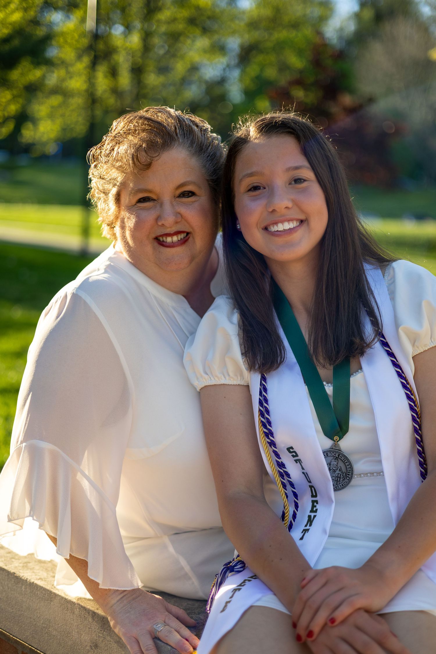 Two women smile together outdoors, younger woman wearing graduation medal and honor cords at Slippery Rock University.