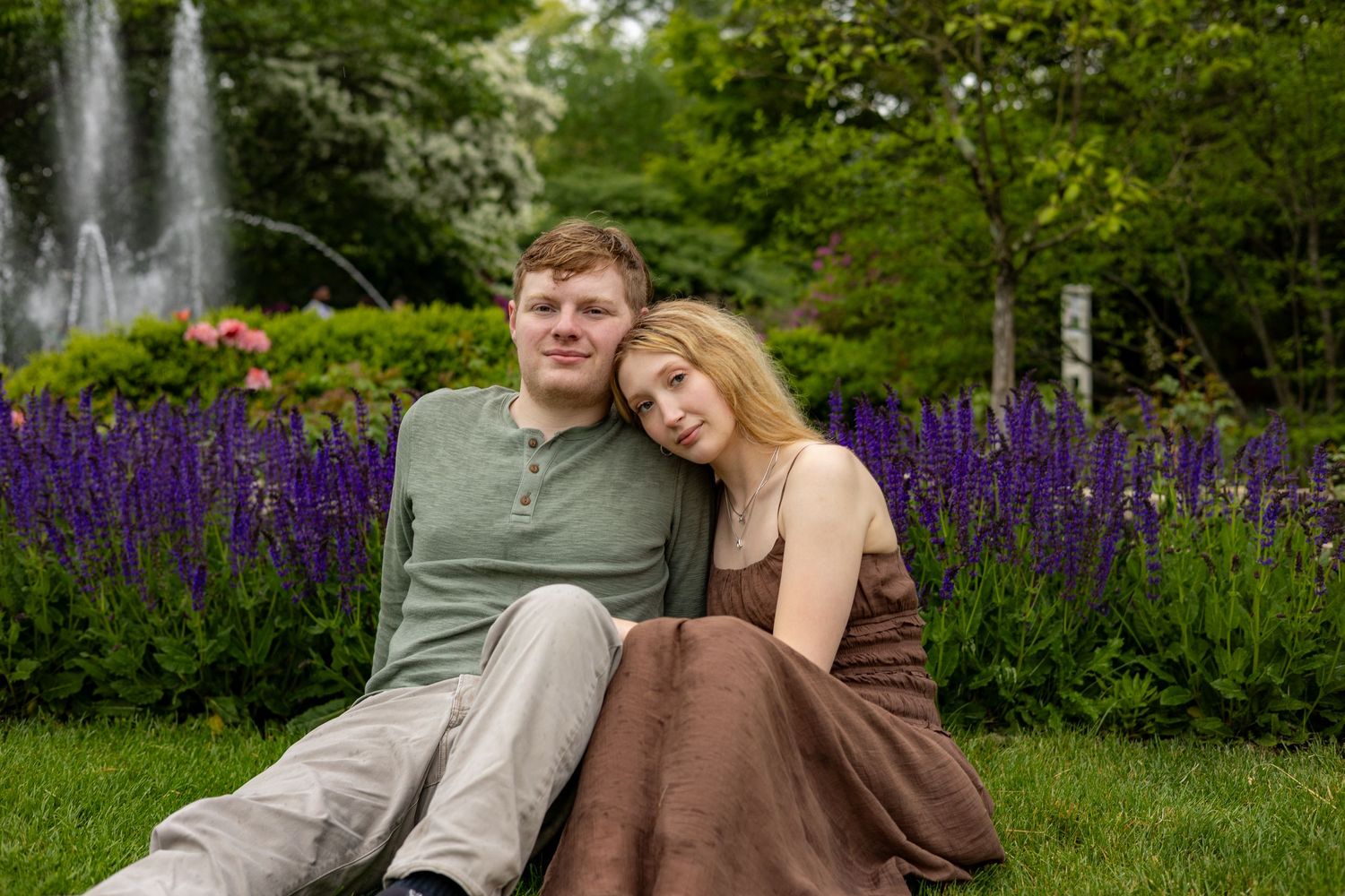 A couple sits together on green grass in front of a fountain and purple flowers.