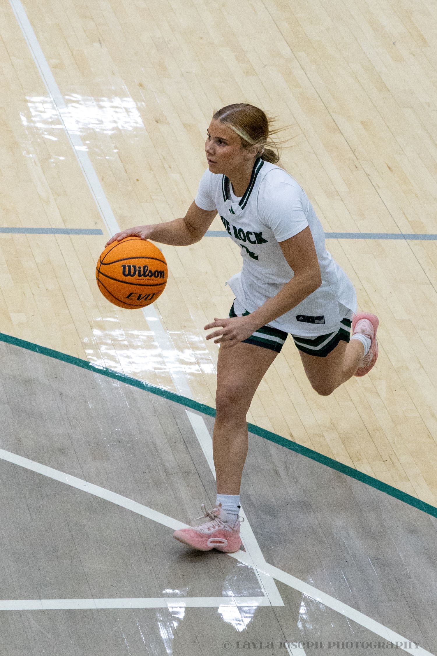 Female basketball player in white The Rock uniform dribbling a Wilson basketball on court.