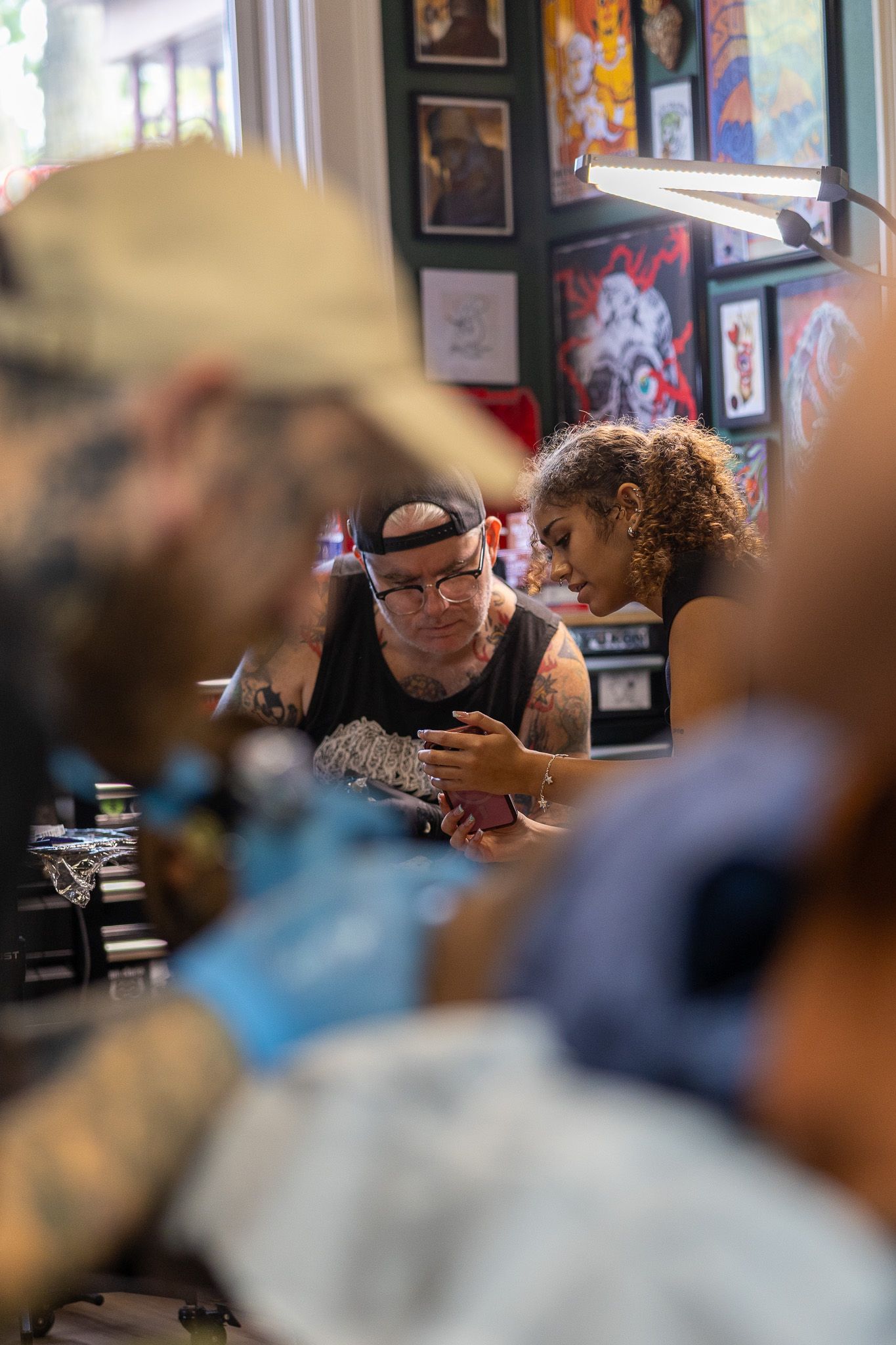 Tattoo artist with cap and tattoos works on a client in a vibrant tattoo shop filled with artwork.