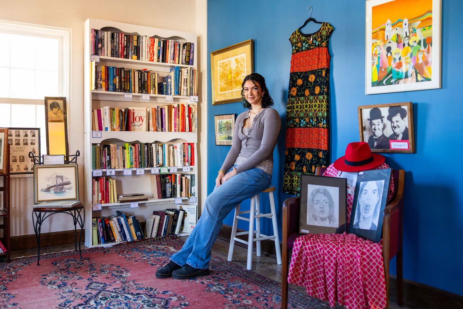 Woman poses in colorful room with bookshelf, vintage dress, artwork, and red hat on chair.