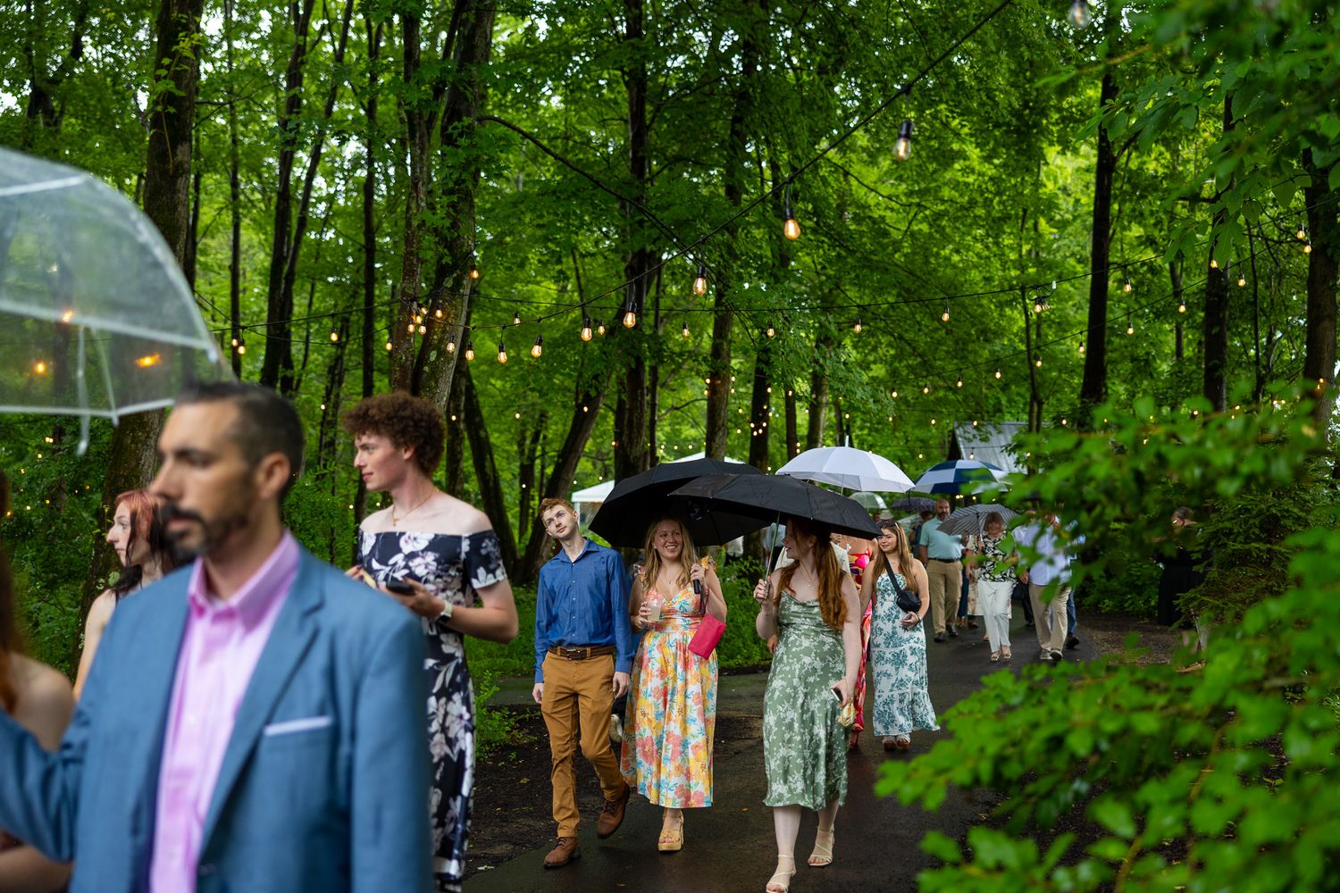 Wedding guests walk through lush green forest path carrying umbrellas in the rain at an outdoor ceremony.