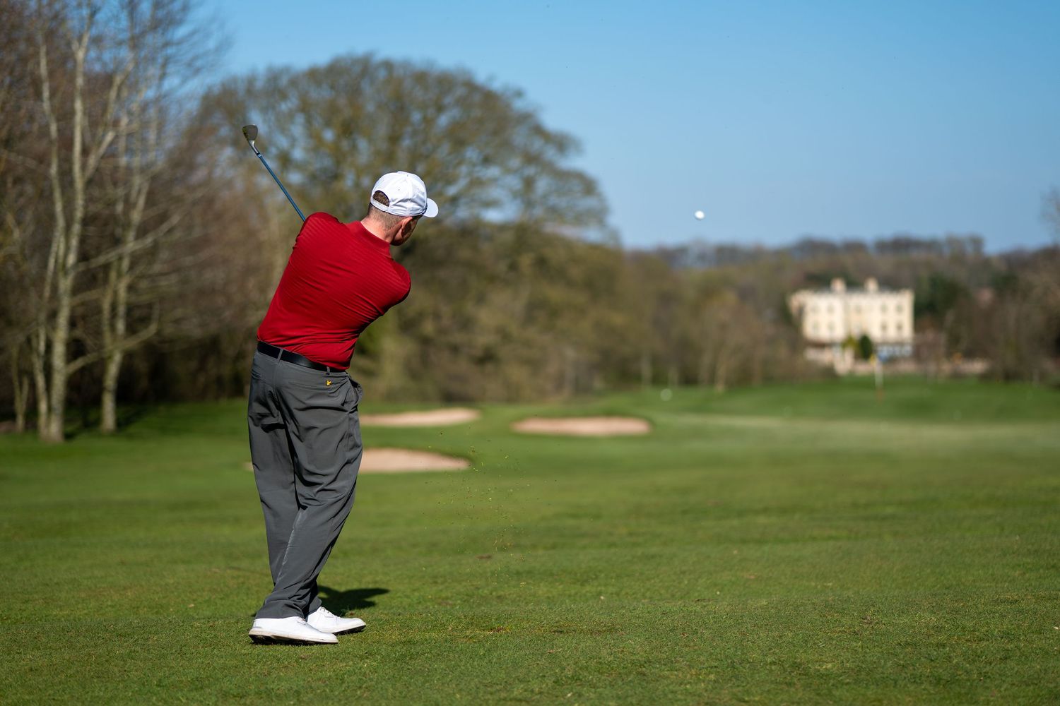 Golfer in red shirt swinging club on lush green fairway with mansion backdrop.