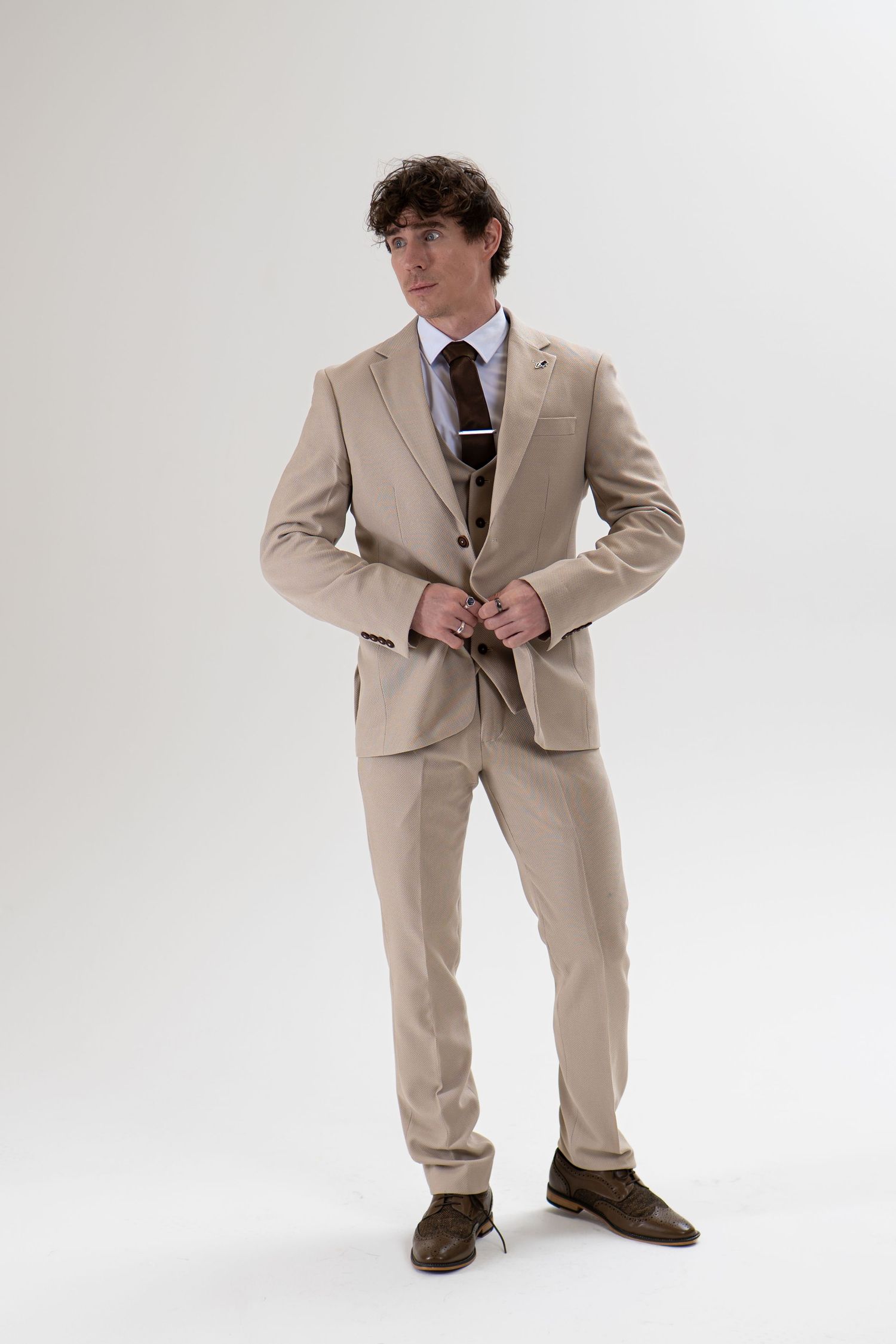 Man in beige three-piece suit fastening jacket button, looking away in studio.