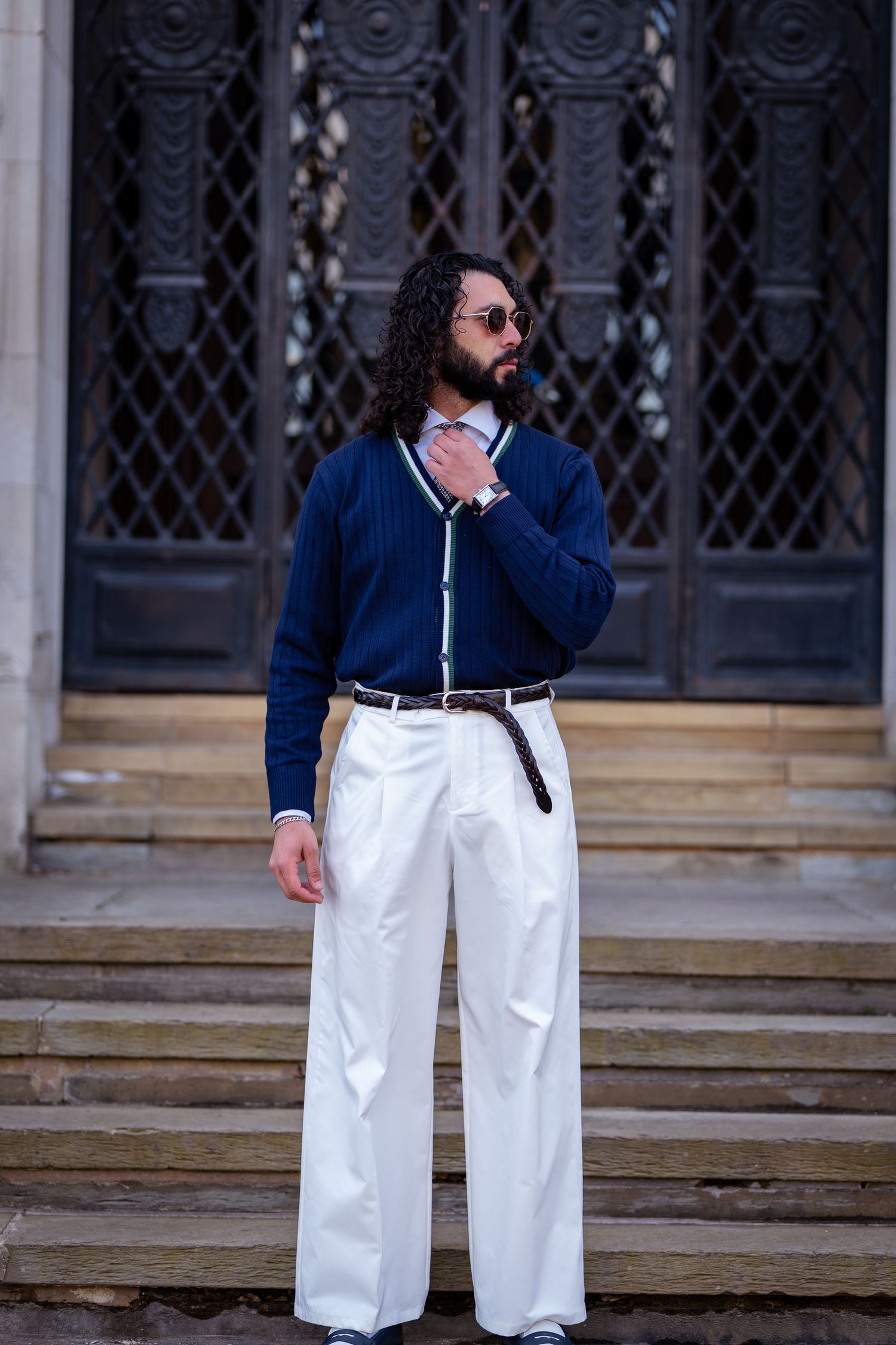 Fashionable man in navy cardigan and wide-leg white trousers standing on steps before ornate dark doors.