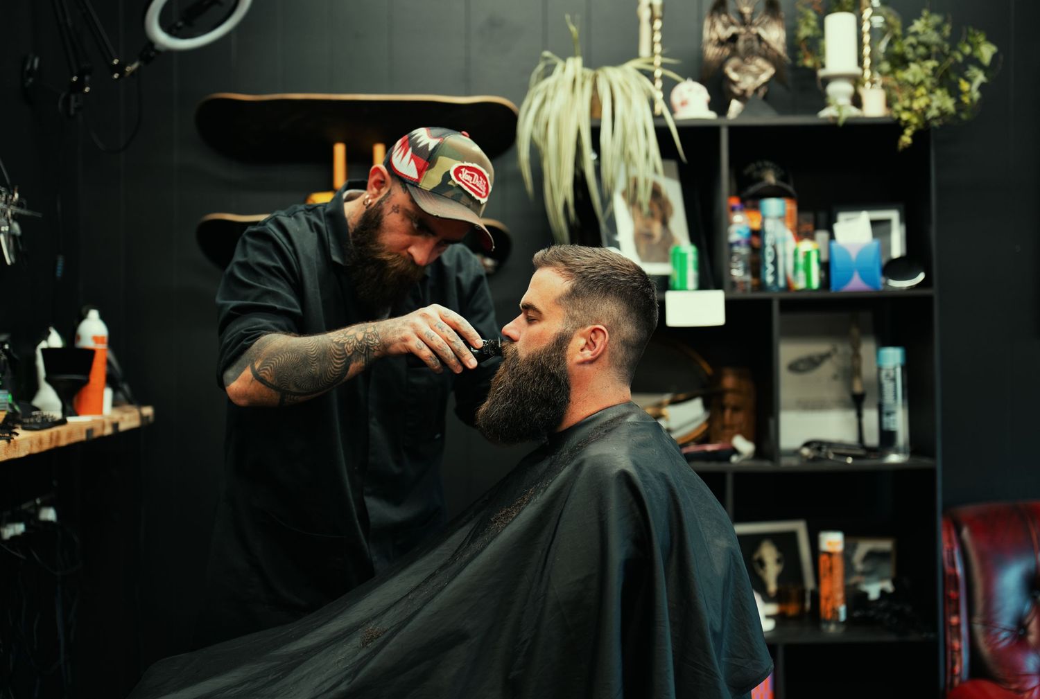 Tattooed barber trimming a bearded man's hair in a stylish dark barbershop with shelves of products.