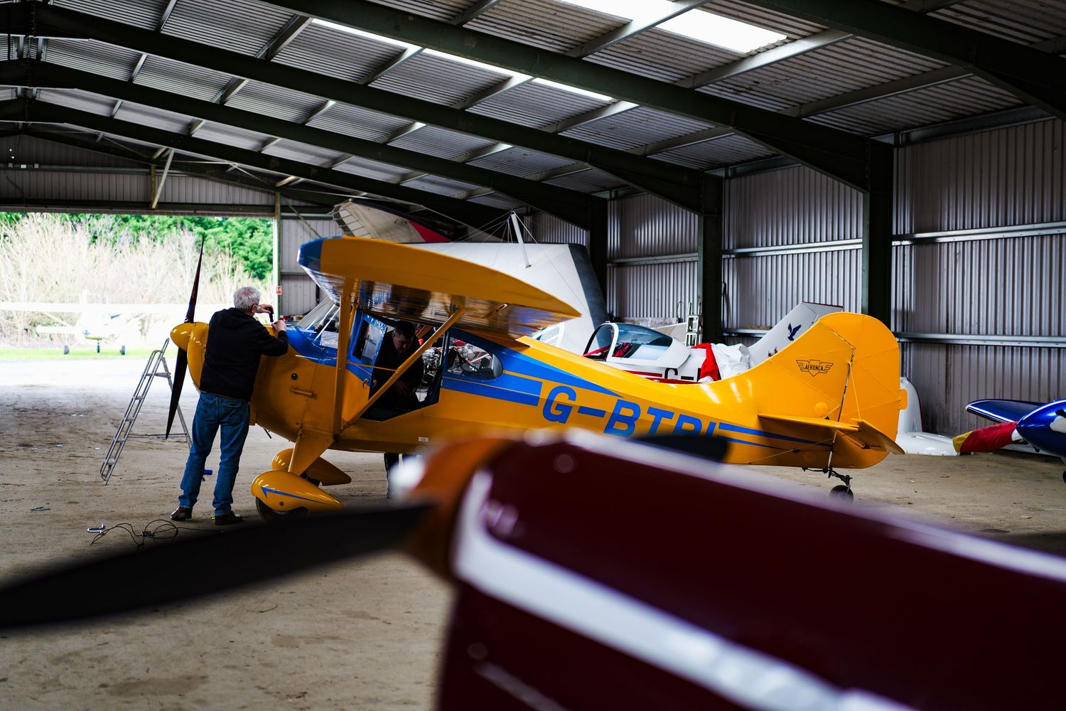 Yellow biplane with registration G-BT inside a spacious aircraft hangar with other planes visible.