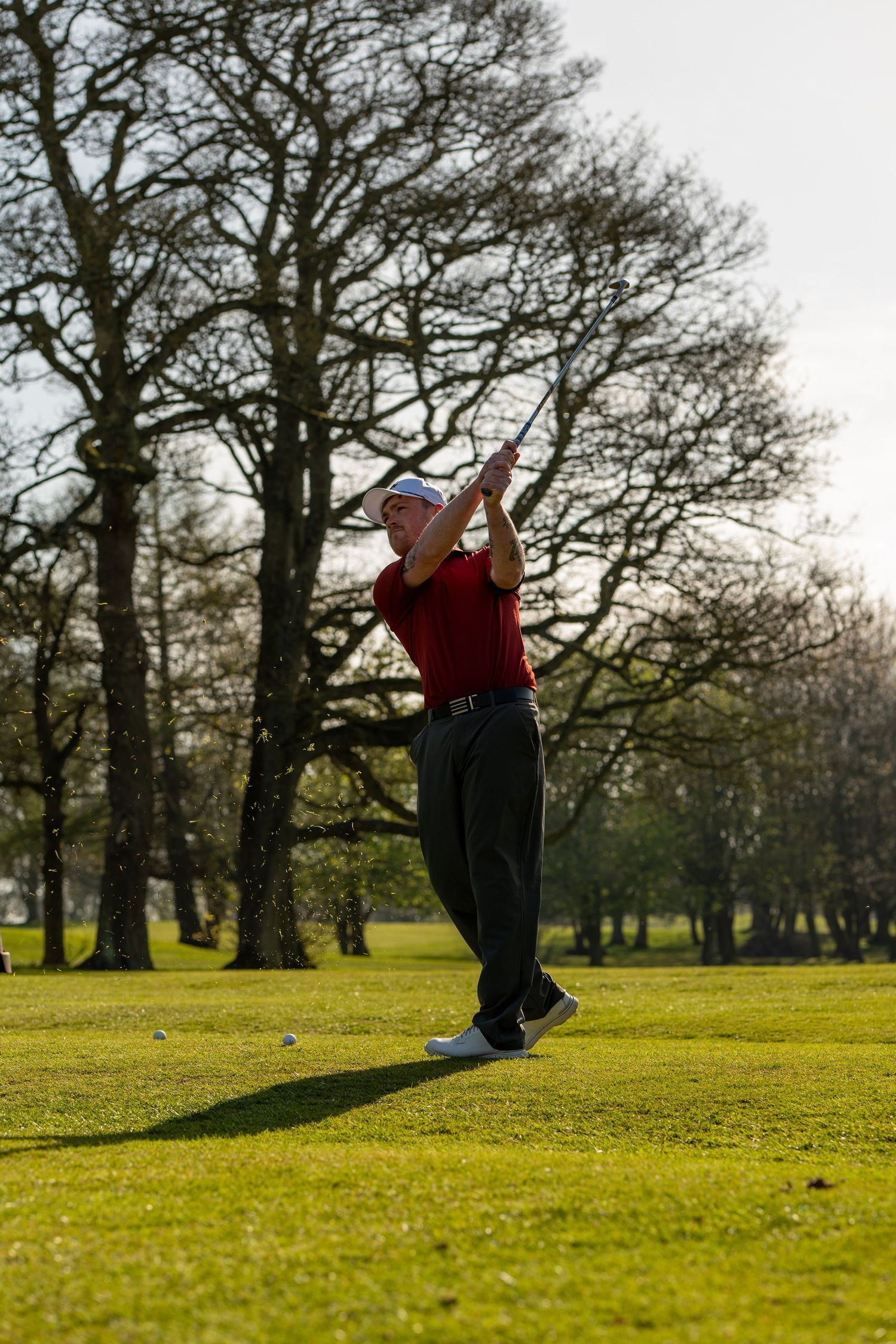 Male golfer in red shirt finishing upward follow-through swing on golf course with tall bare trees behind.