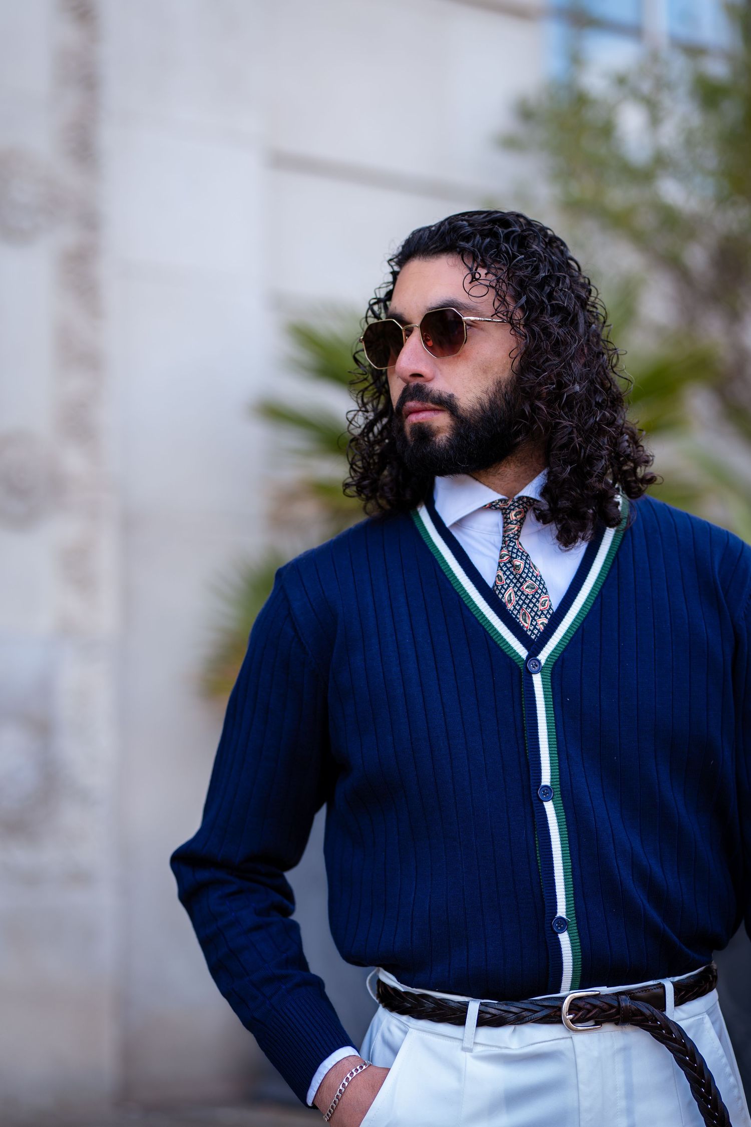 Stylish bearded man in navy cardigan with green trim wearing round sunglasses and patterned tie outdoors.